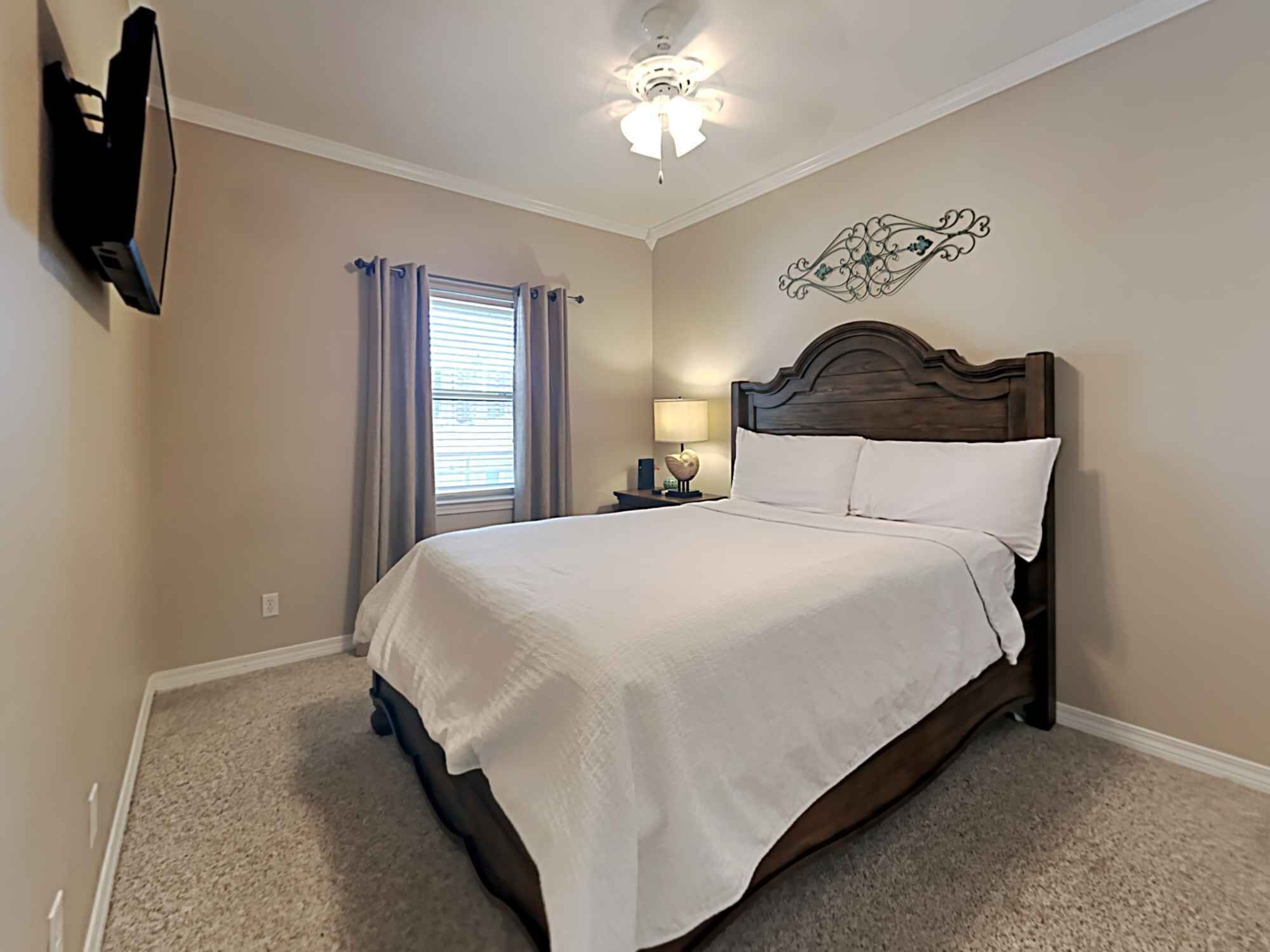 A bedroom with a bed, a ceiling fan, a wall-mounted TV, a window with curtains, and a decorative wall piece.