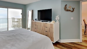 Een slaapkamer met een bed, een televisie op een houten kast, een plafondventilator en een balkon met uitzicht op de oceaan.