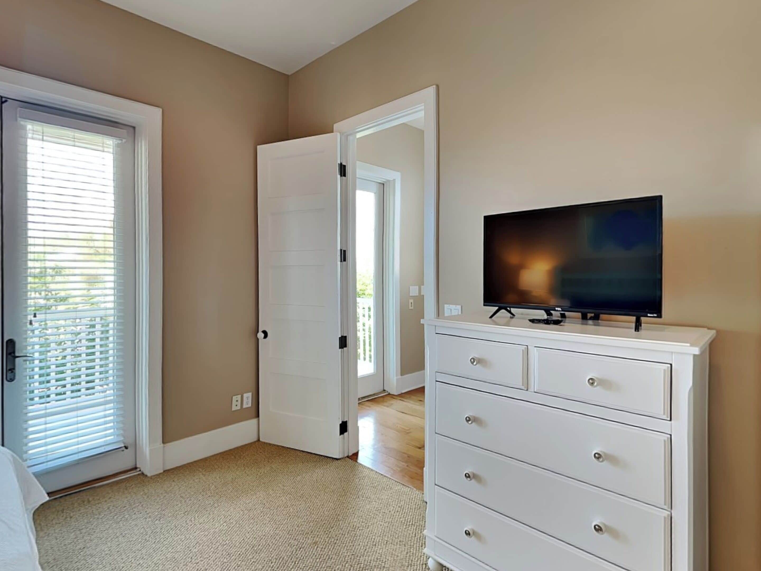 Una habitación con una televisión sobre un tocador blanco, una puerta y una ventana con persianas.