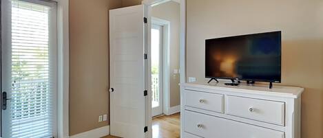 Una habitación con una televisión sobre un tocador blanco, una puerta y una ventana con persianas.