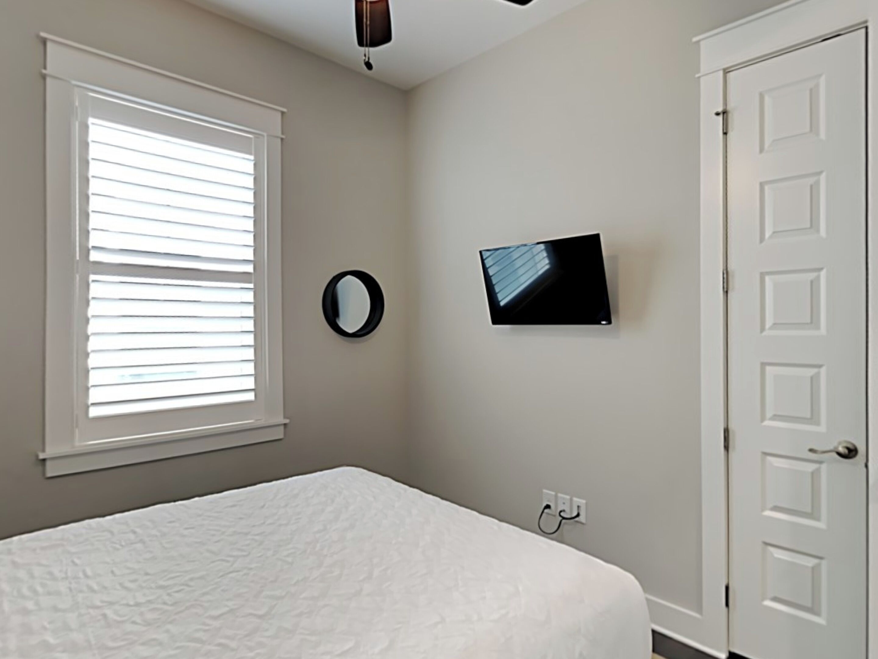 A bedroom with a bed, a TV mounted on the wall, a window with blinds, and a door.