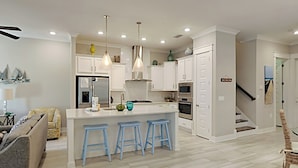 A modern kitchen with white cabinets, a central island with blue bar stools, and a dining area with a grey sofa and patterned armchair. - Prominence North #94A - Happy Ours by Southern Vacation Rentals (Santa Rosa Beach)