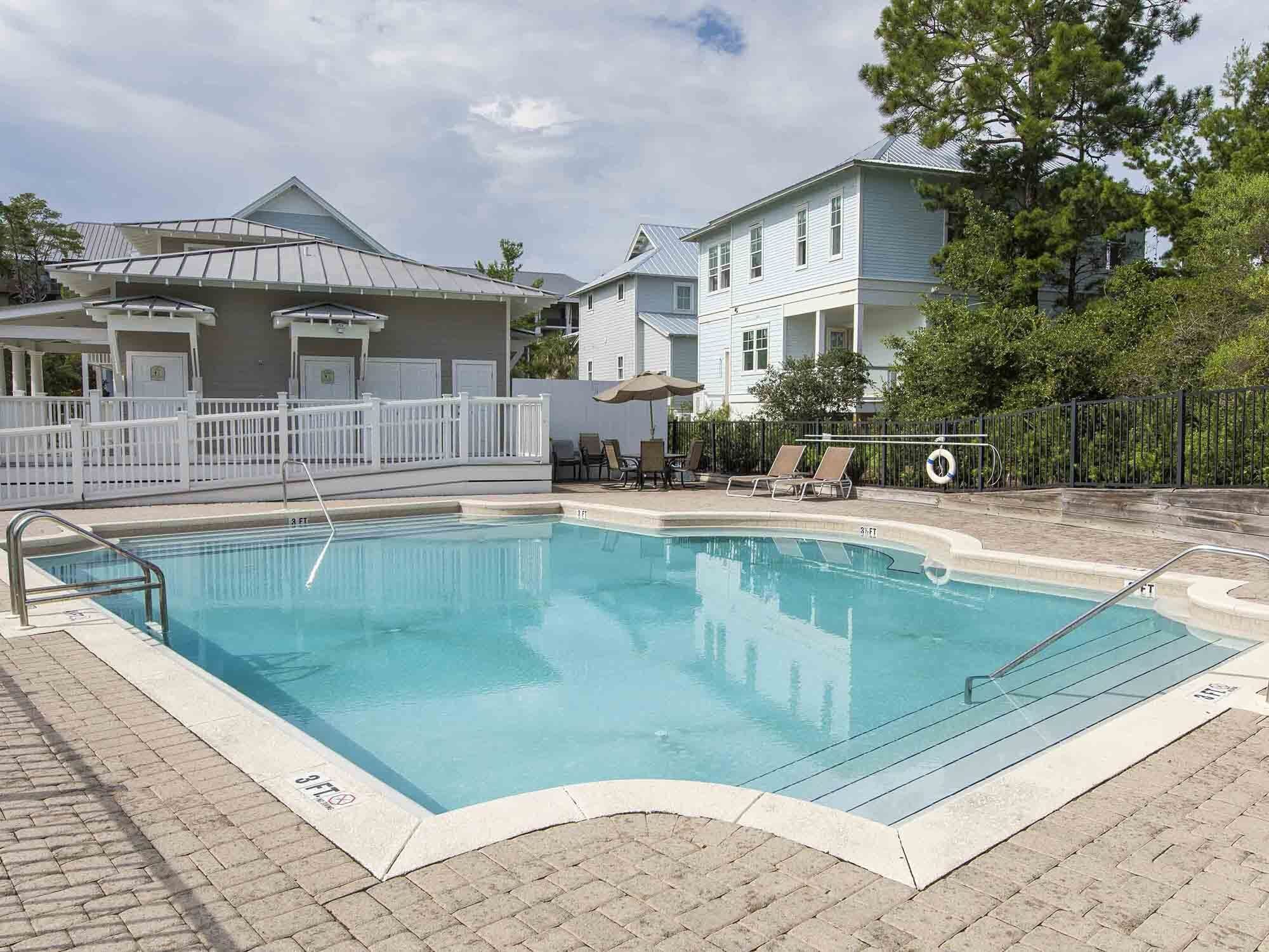 A swimming pool with a white deck, lounge chairs, and a fenced area.