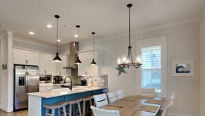 A modern kitchen with white cabinets, a stainless steel refrigerator, a central island with bar stools, pendant lighting, and a dining area with a wooden table and white chairs. - Starfish Sanctuary by Southern Vacation Rentals (Santa Rosa Beach)