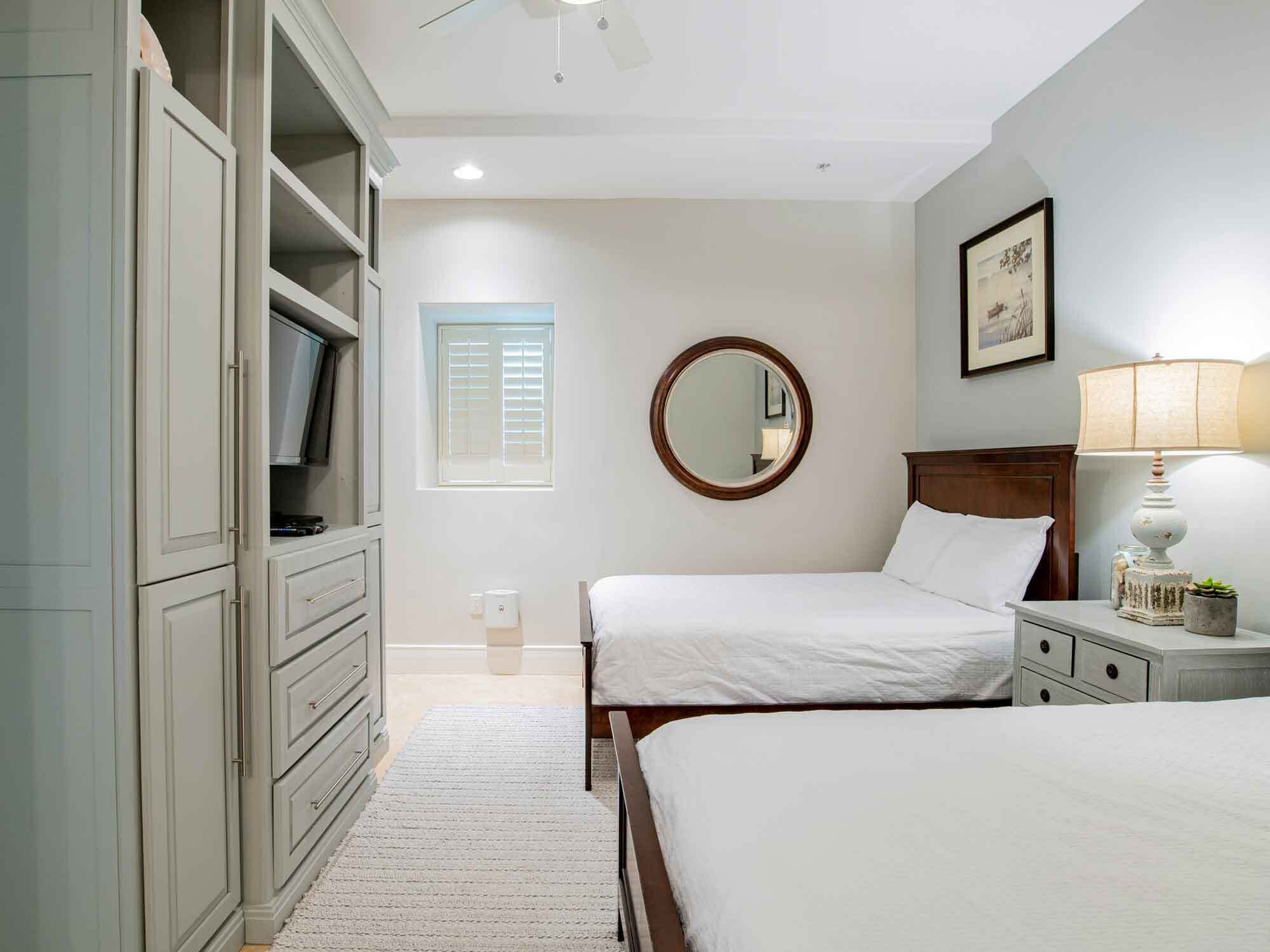 A bedroom with two beds, a wardrobe, a round mirror, a bedside table with a lamp, and a window with blinds.