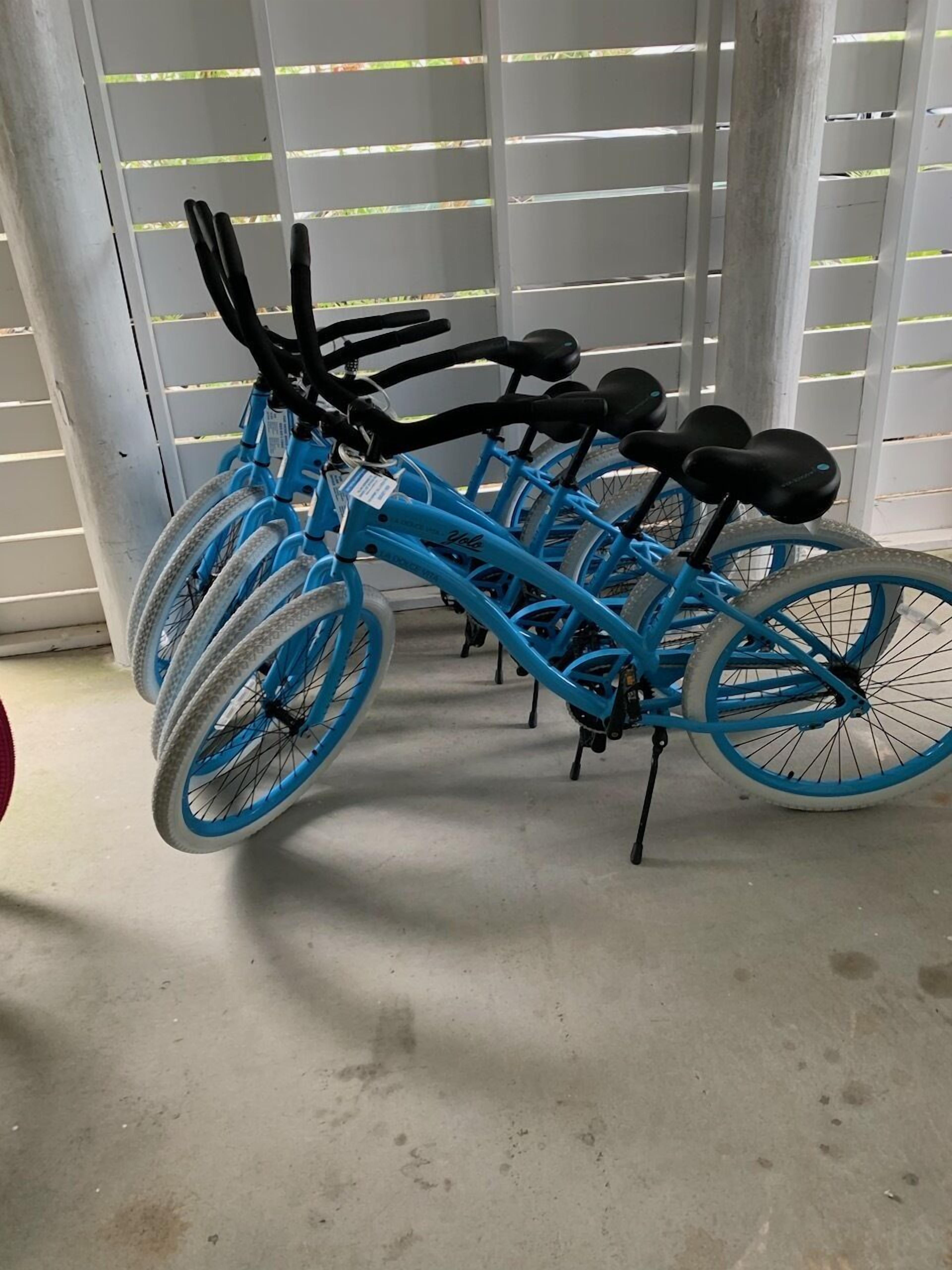 A row of blue bicycles parked indoors, with a concrete floor and a white fence in the background.
