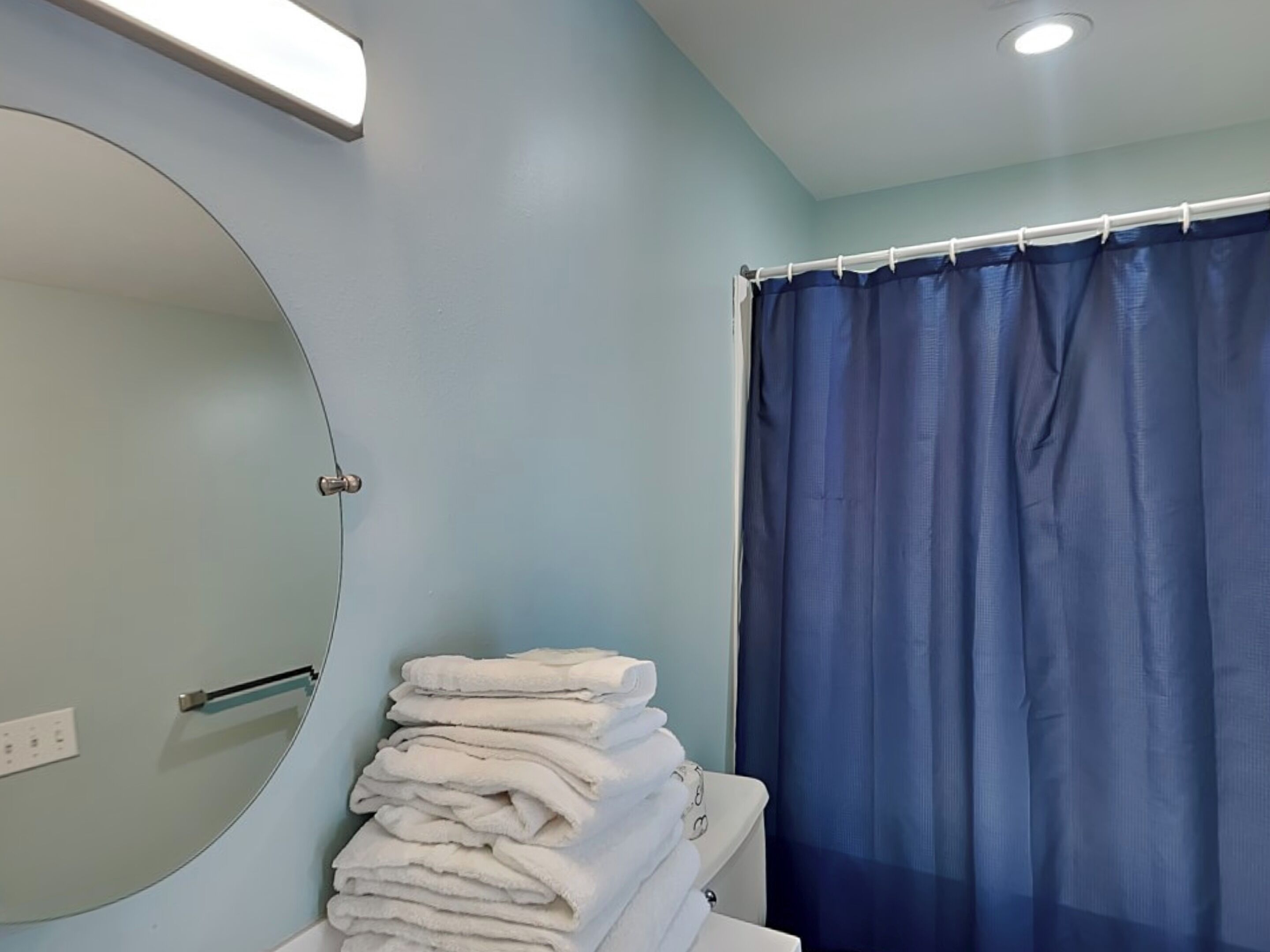 A bathroom with a round mirror, a stack of white towels, and a blue shower curtain.