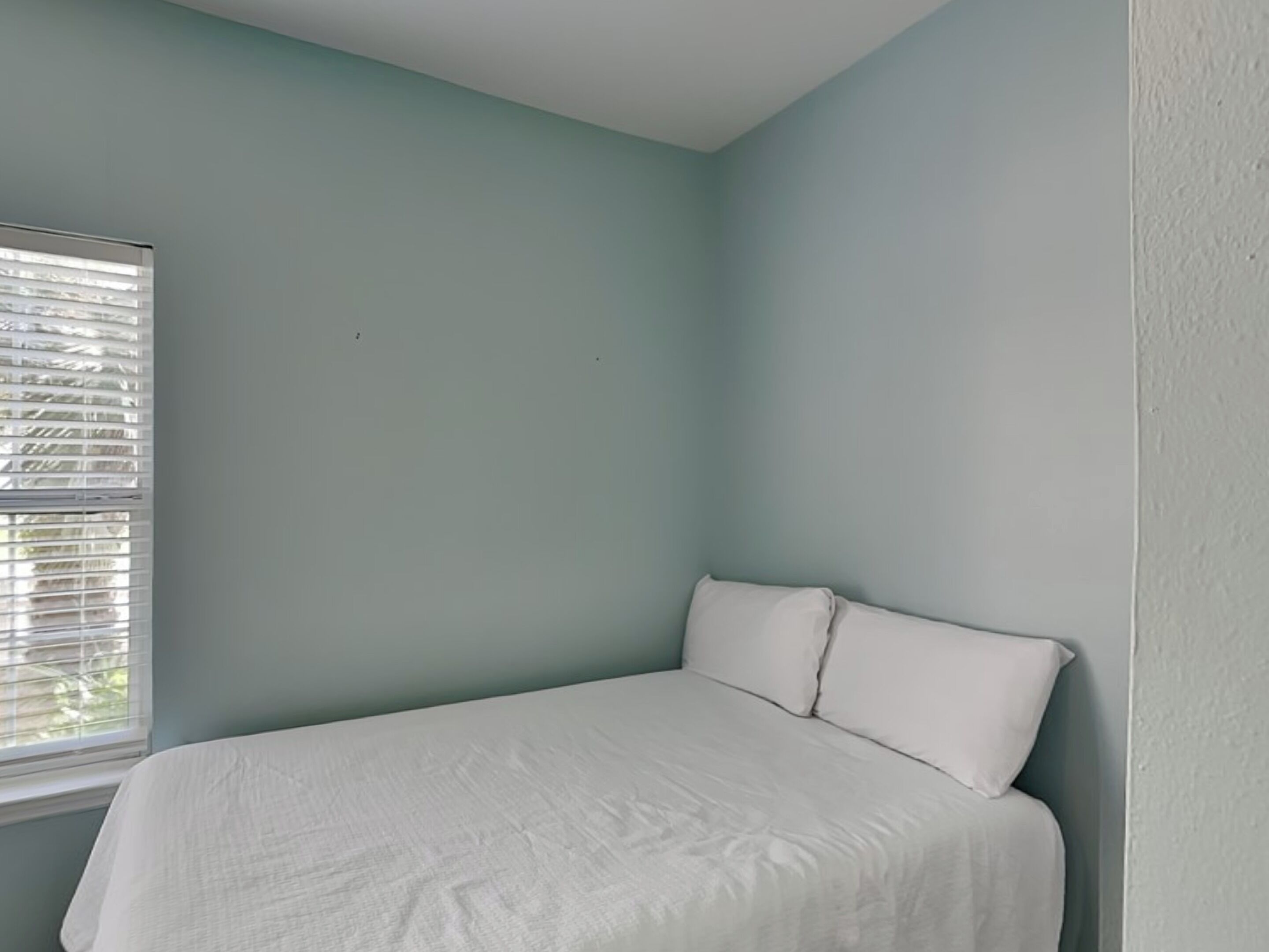 Une chambre avec un lit simple, des murs bleu clair et une fenêtre avec des stores.