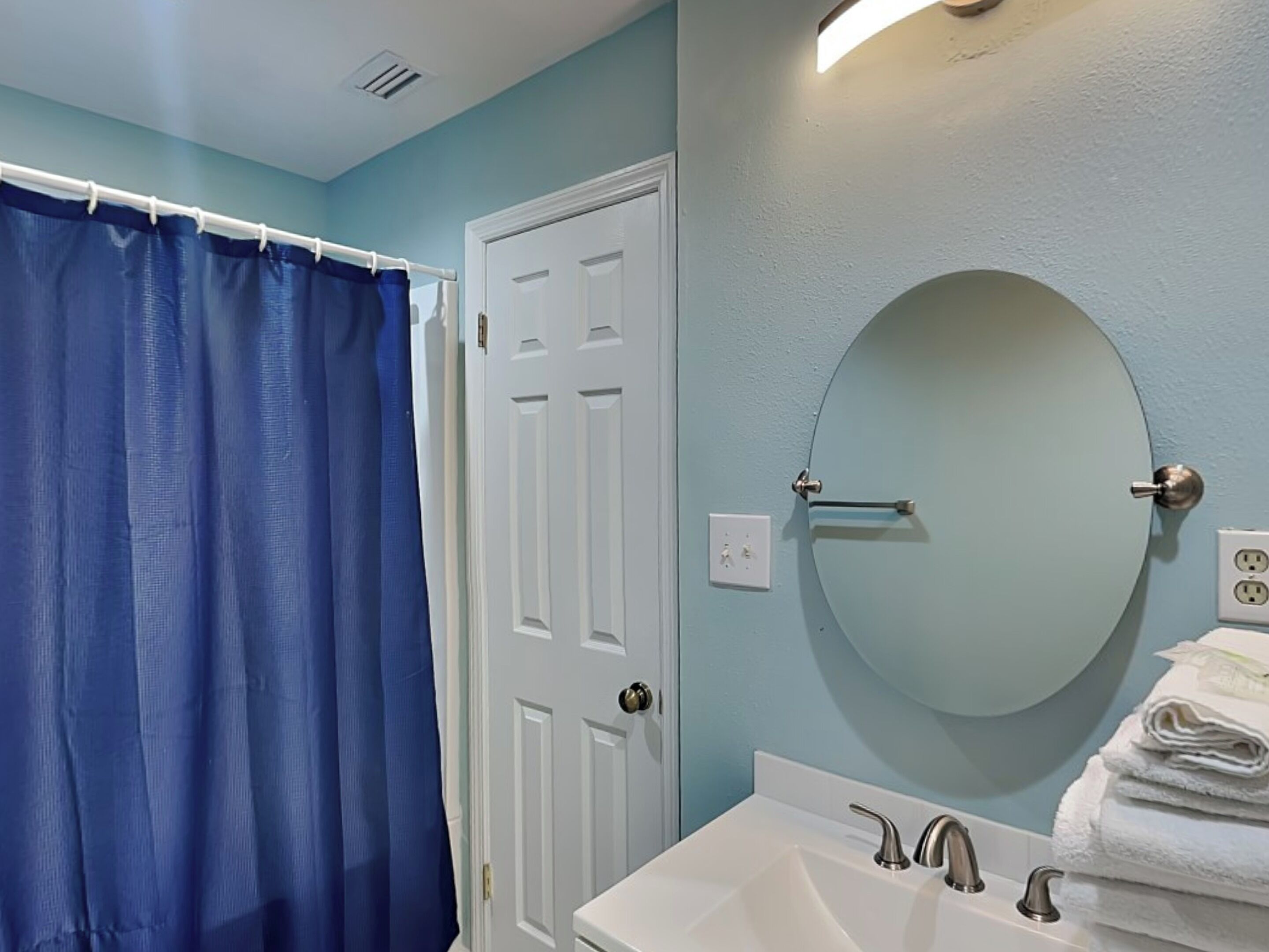 Une salle de bain avec un rideau de douche bleu, une porte blanche, un miroir rond et un lavabo avec deux robinets.