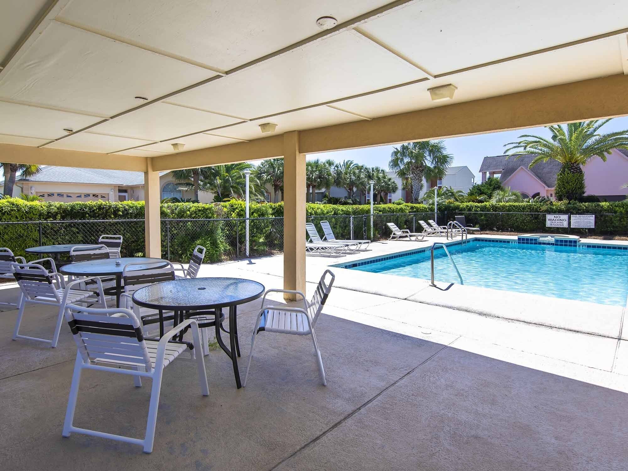 A covered pool area with lounge chairs and tables, a swimming pool, and a view of palm trees and buildings in the background.