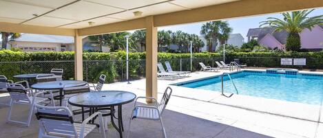 A covered pool area with lounge chairs and tables, a swimming pool, and a view of palm trees and buildings in the background.