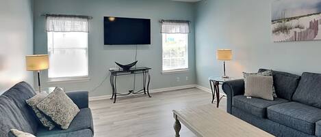 Una sala de estar con dos sofás, una mesa de centro, un televisor y dos ventanas con cortinas.
