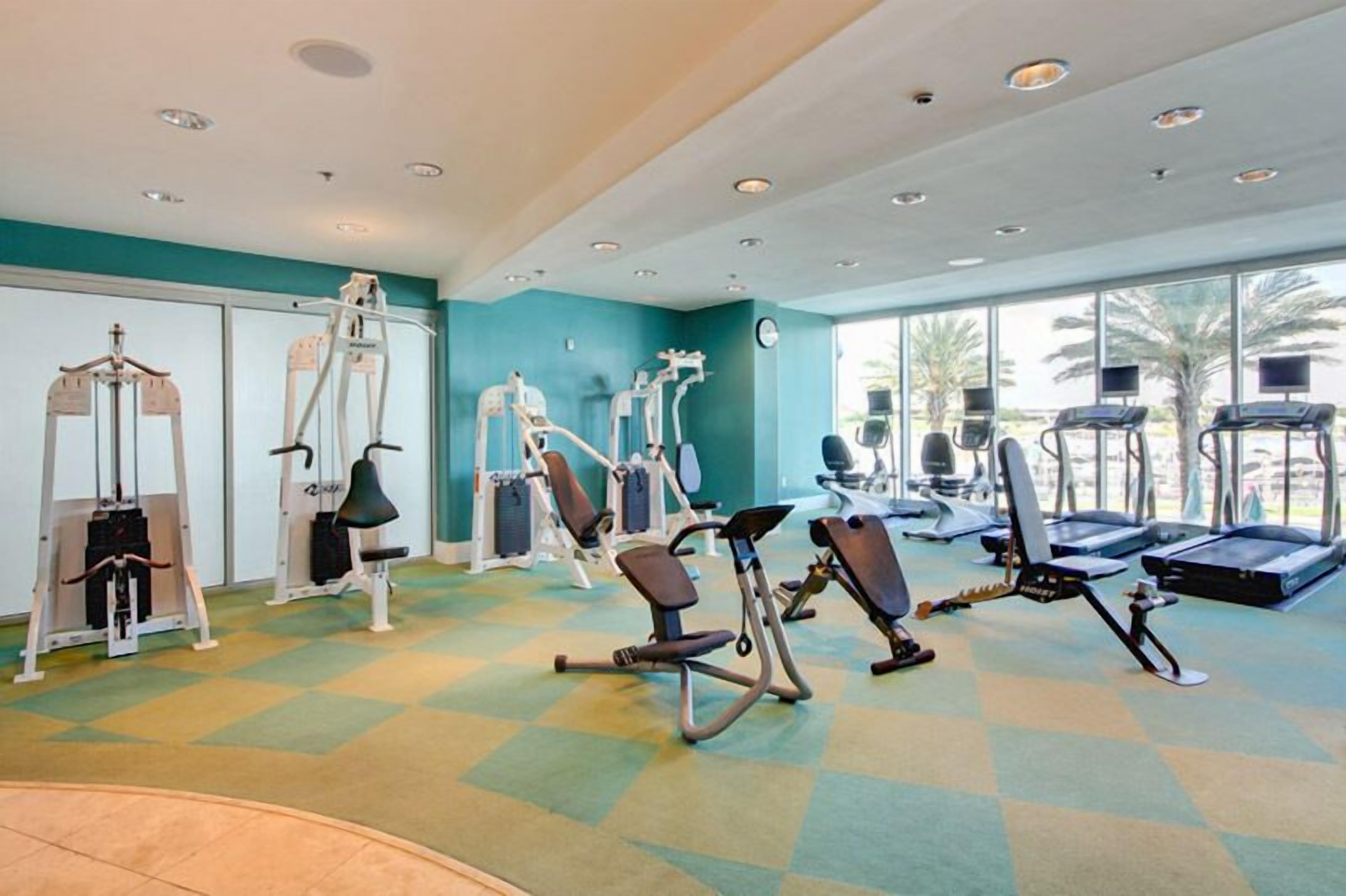 Ein Fitnessraum mit unterschiedlichen Trainingsgeräten, großen Fenstern und einer Türkiswand.