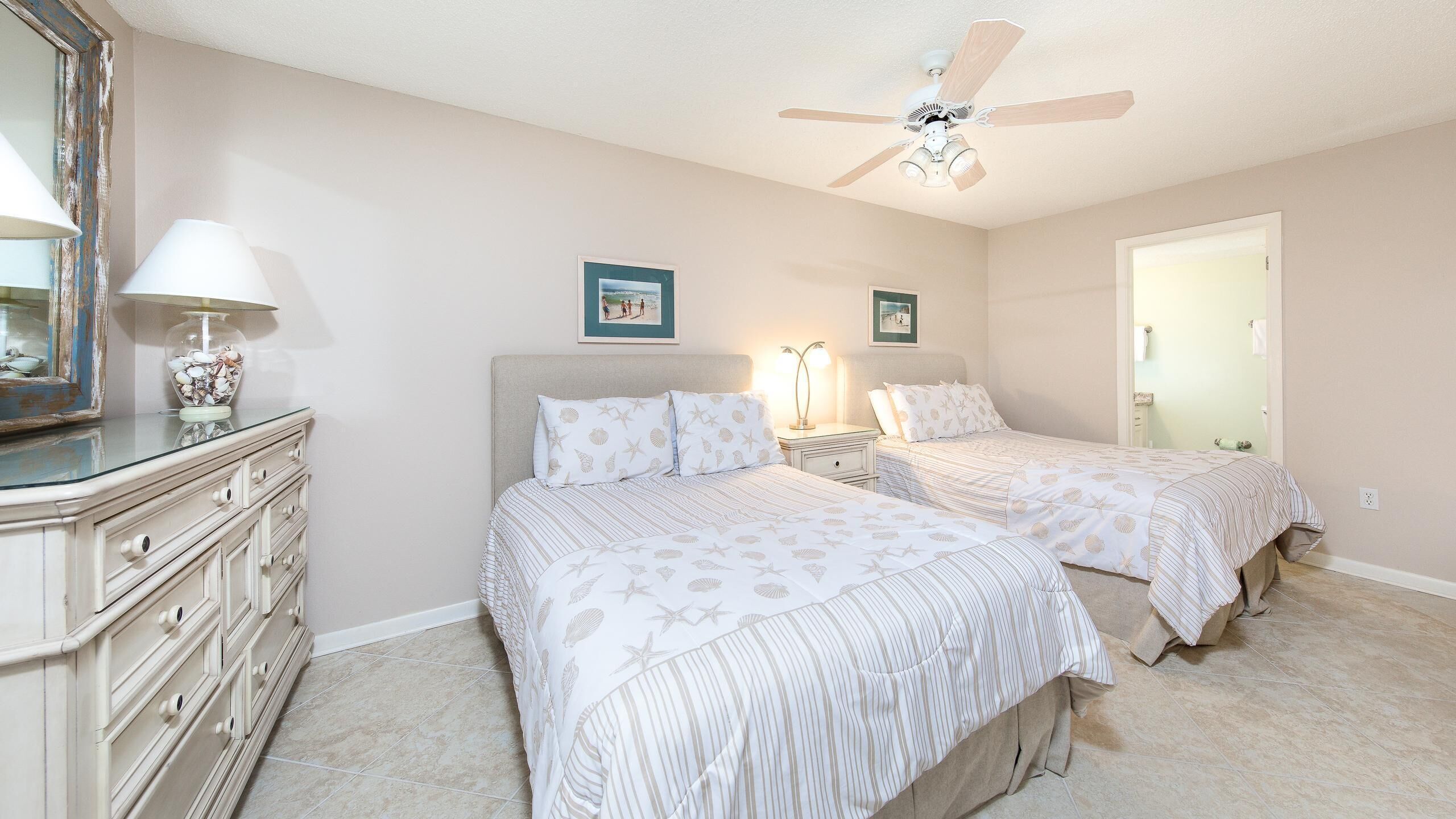 A bedroom with two beds, a dresser, a ceiling fan, and a door leading to another room.