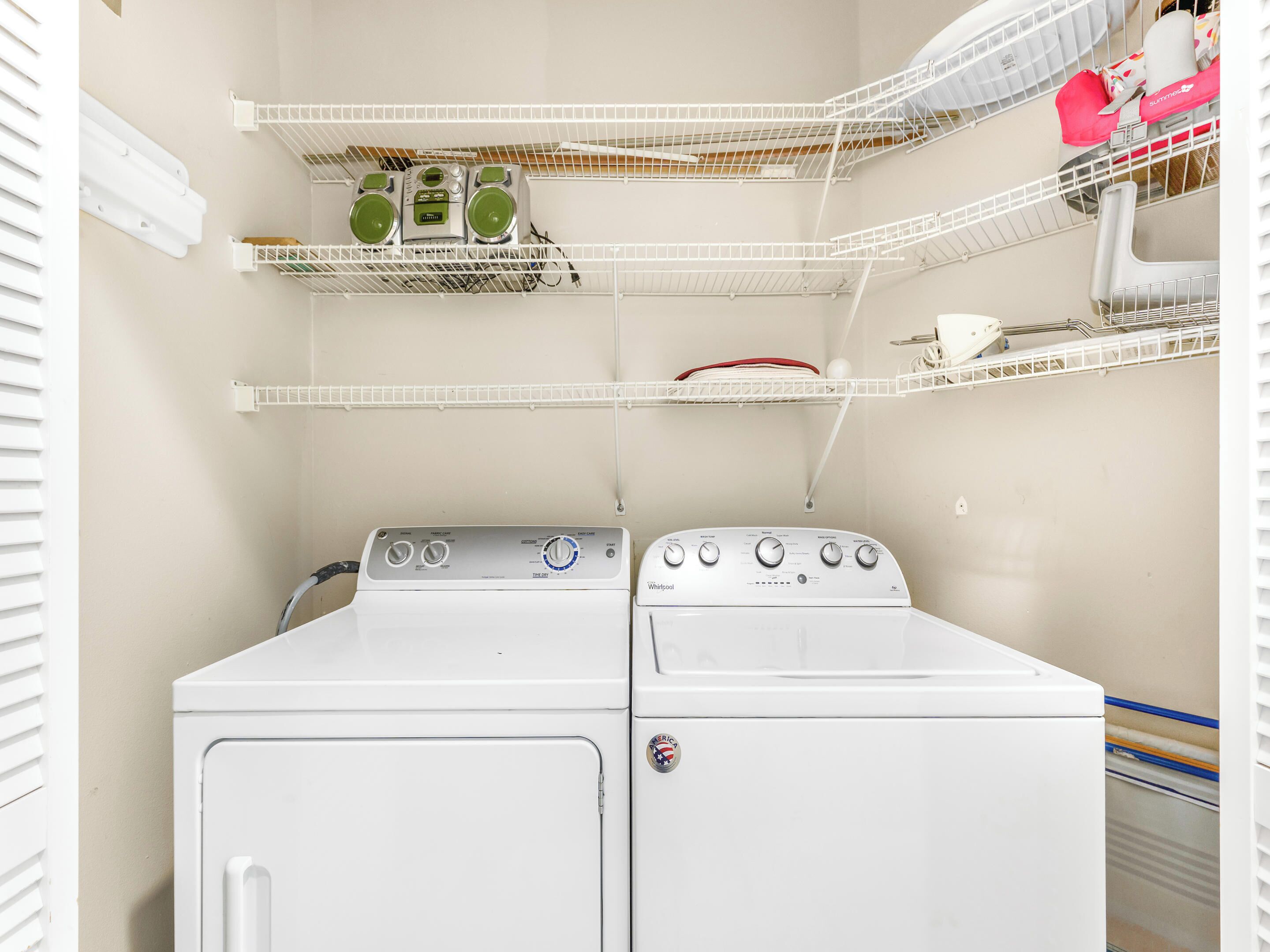 A laundry area with two stacked washers and dryers, white shelving units with various items, and a window with blinds.