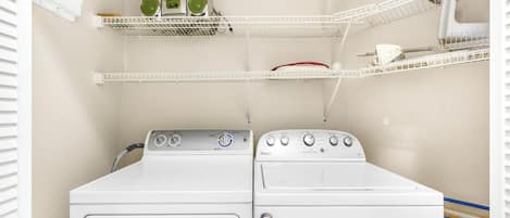 A laundry area with two stacked washers and dryers, white shelving units with various items, and a window with blinds.