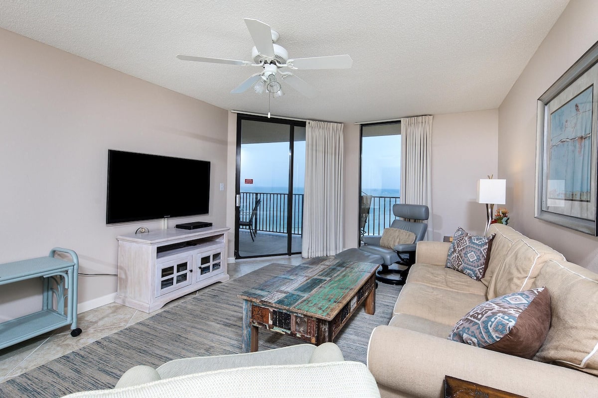 A hotel room with a flat-screen TV, a ceiling fan, a sofa, a coffee table, and a balcony with a view.