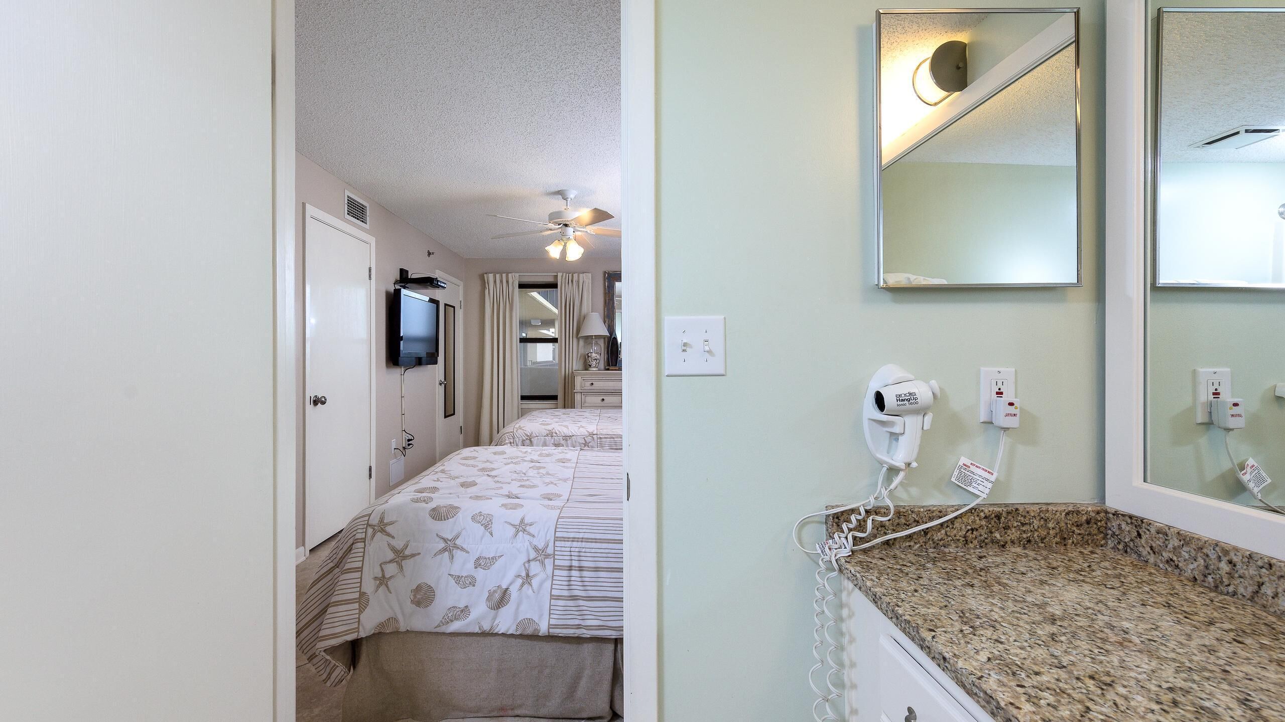 A hotel room with a bed, a television, a ceiling fan, and a bathroom with a sink and mirror.