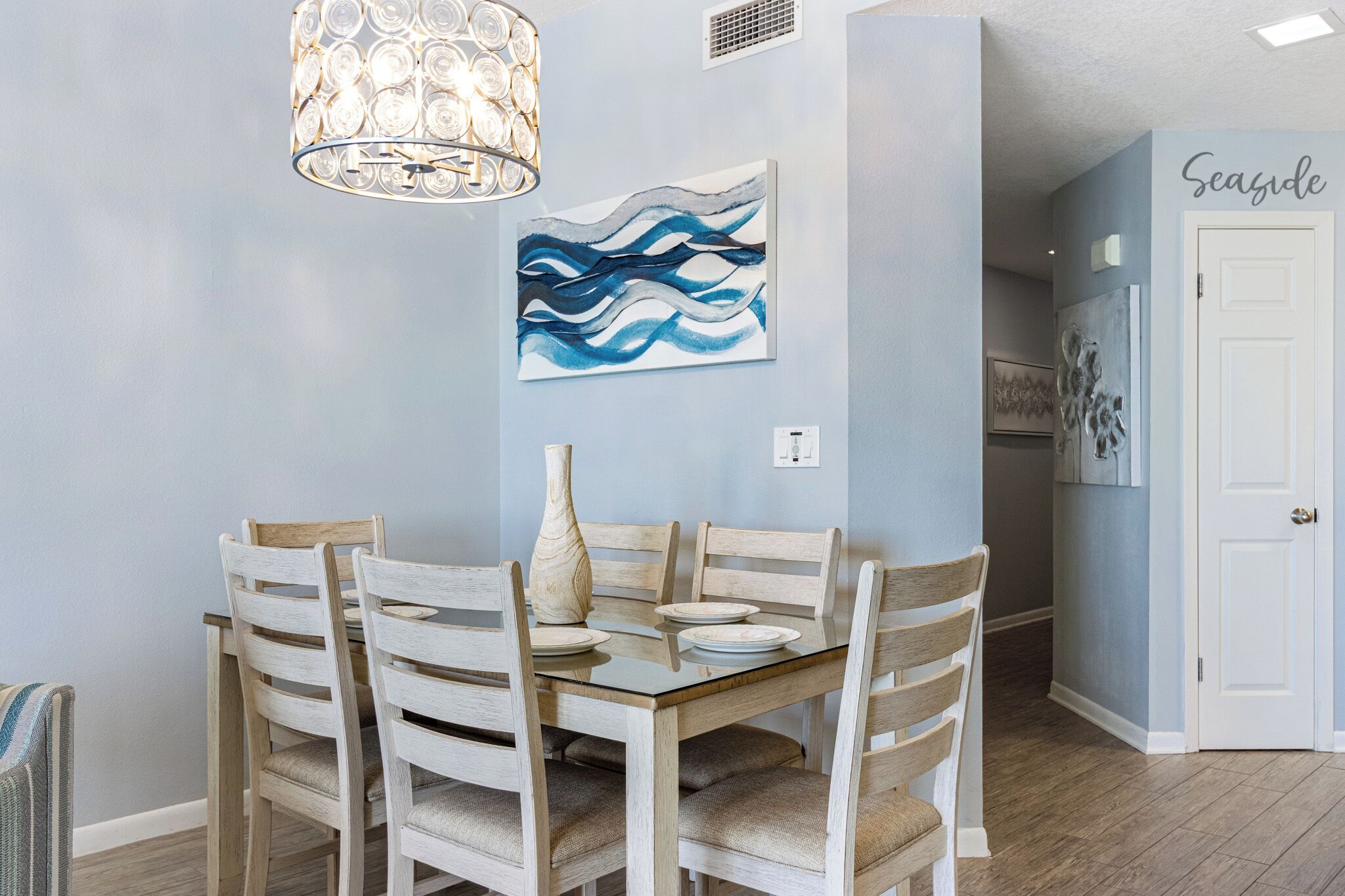 A dining area with a wooden table and chairs, a vase, and a blue abstract painting on the wall.
