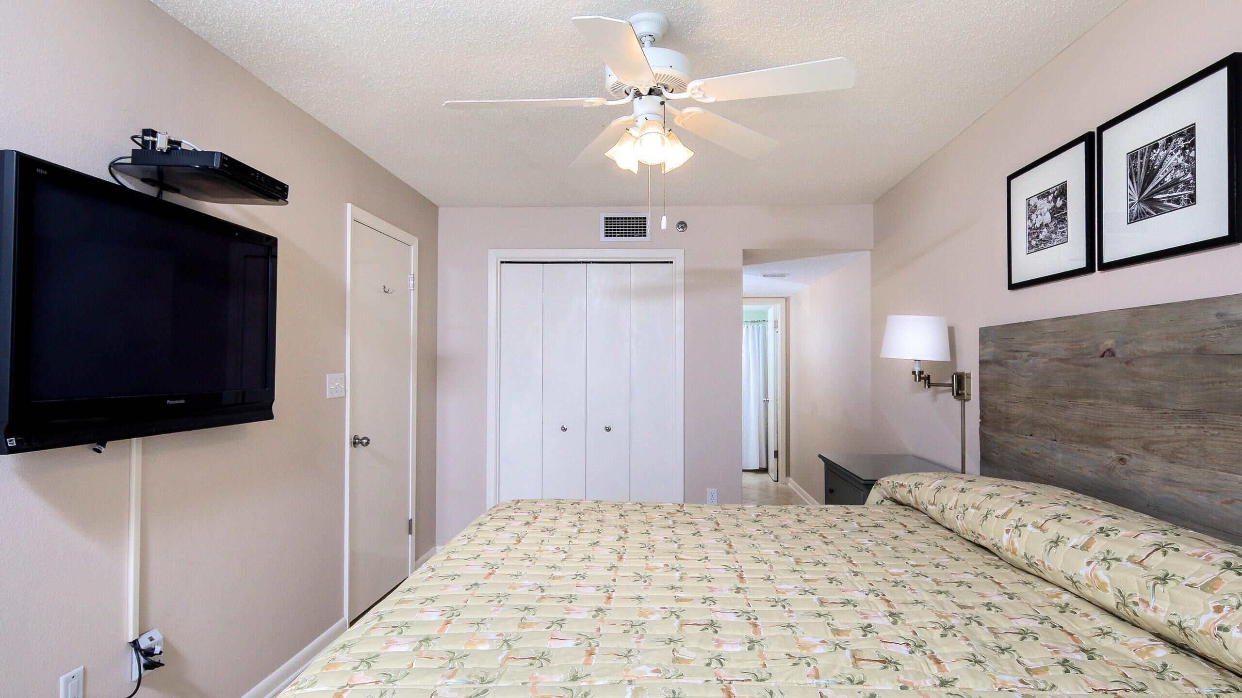 A hotel room with a bed, a ceiling fan, a flat-screen TV mounted on the wall, and two framed pictures on the wall.