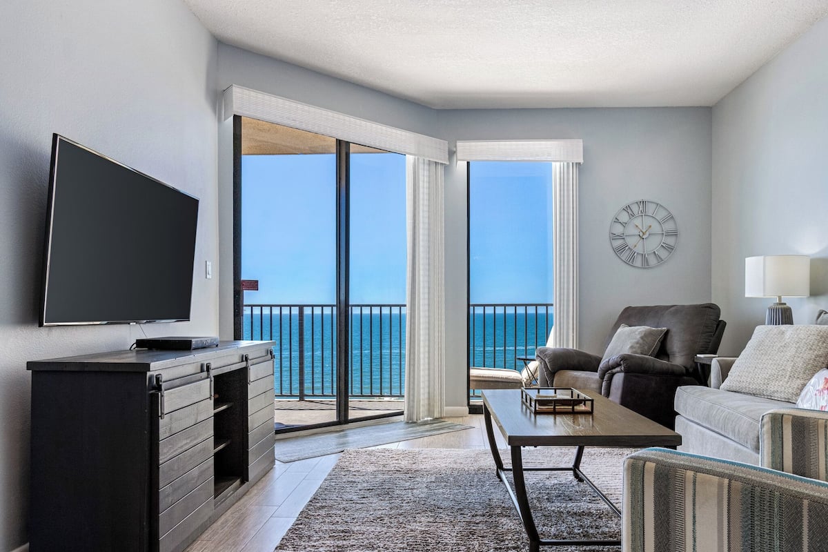 A living room with a large TV, a sofa, a coffee table, and a view of the ocean through large sliding doors.
