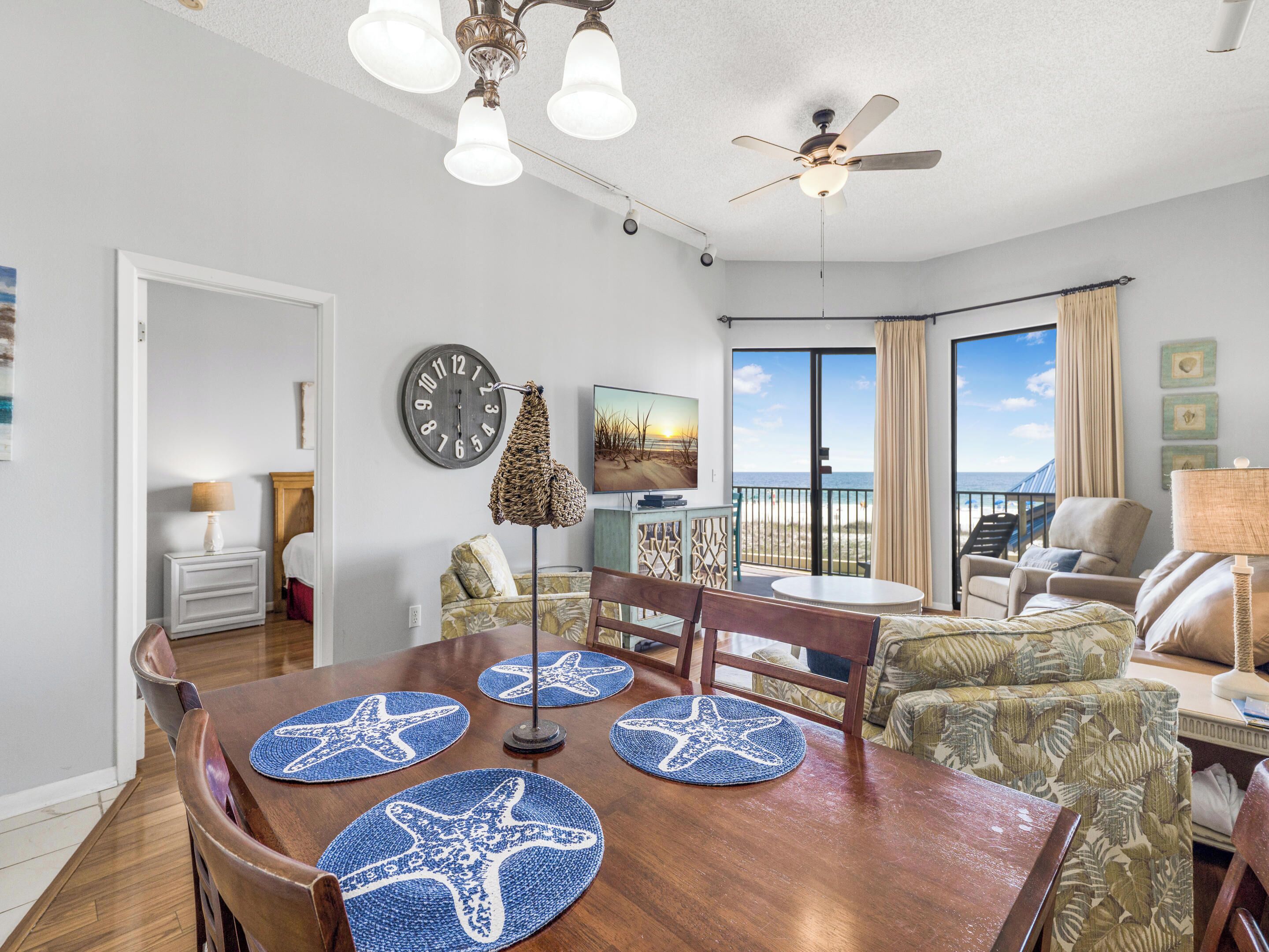 A spacious living room with a wooden dining table, starfish placemats, a large clock, and a view of the ocean through the sliding glass doors.