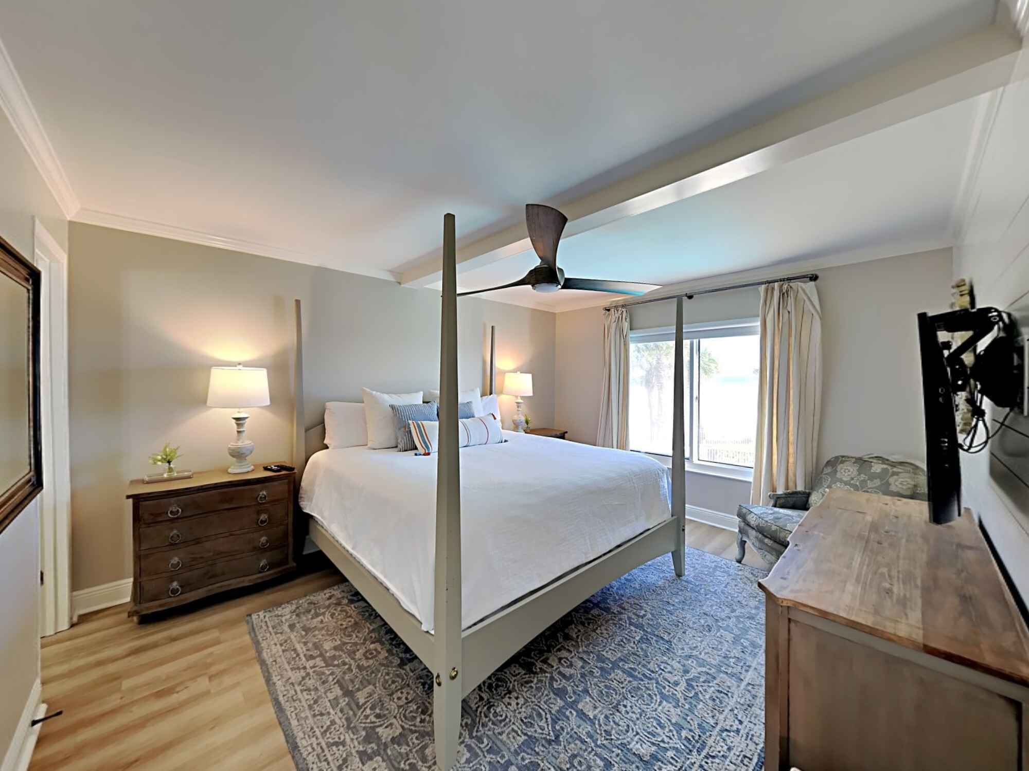 A bedroom with a four-poster bed, a ceiling fan, a nightstand with a lamp, a dresser, and a large window with curtains.