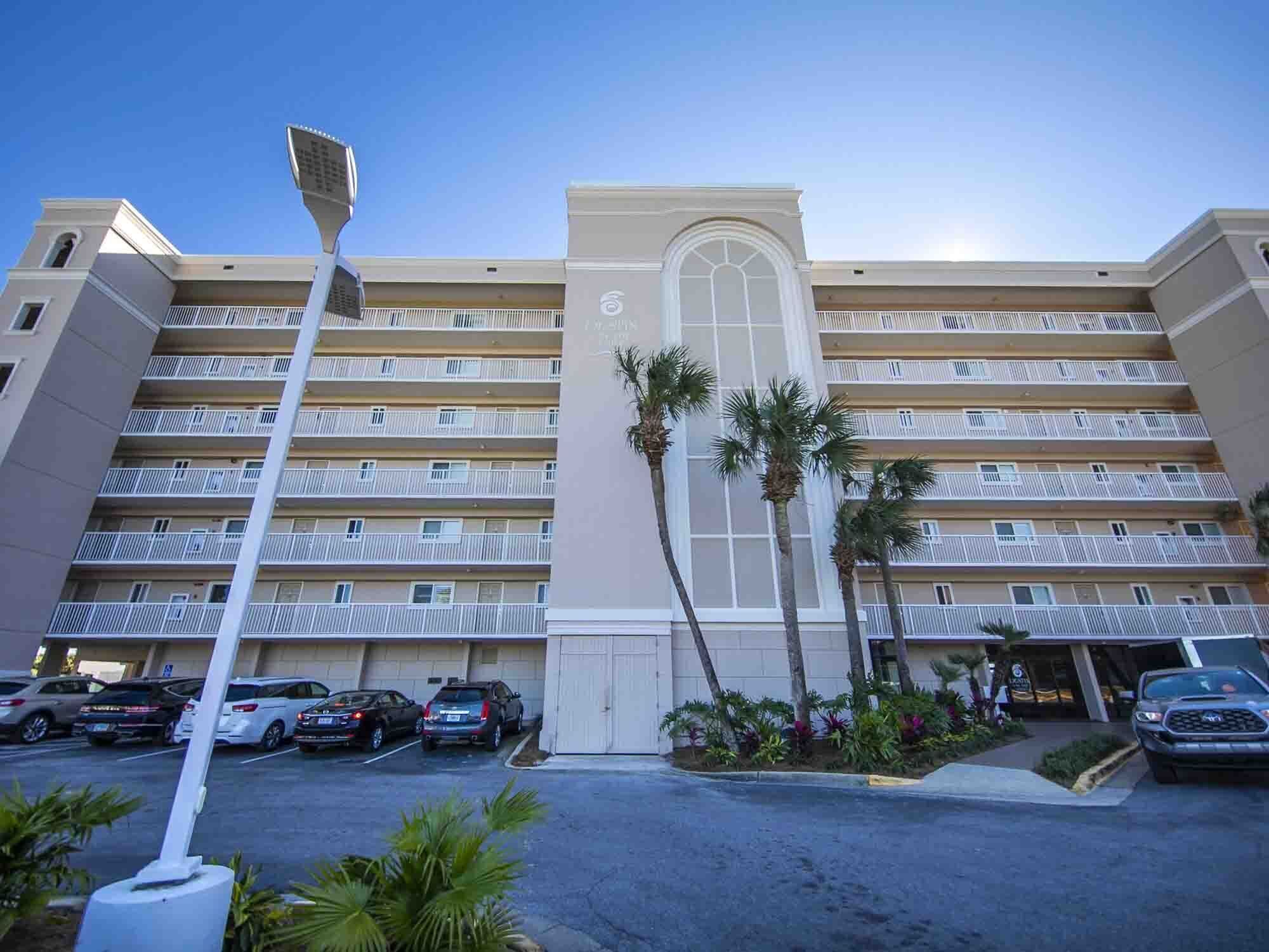 A multi-story hotel with a central entrance, balconies on each floor, and a parking lot with several cars.
