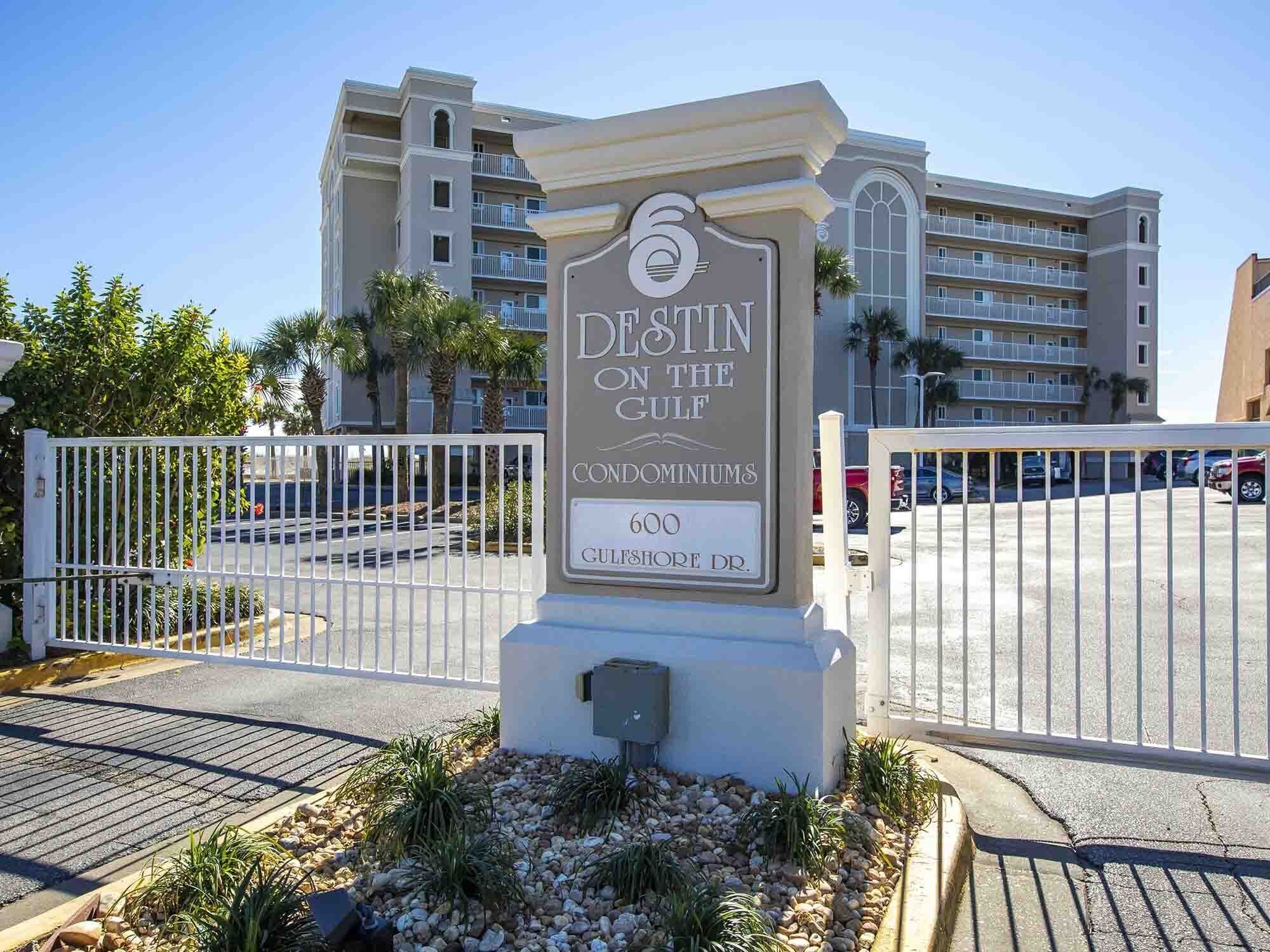 A sign for 'DESTIN ON THE GULF condominiums' with a view of the parking area and buildings in the background.
