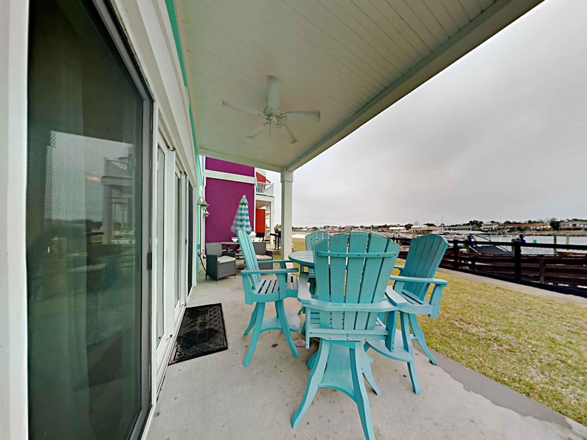 A covered outdoor seating area with turquoise Adirondack chairs and a table, overlooking a waterfront.