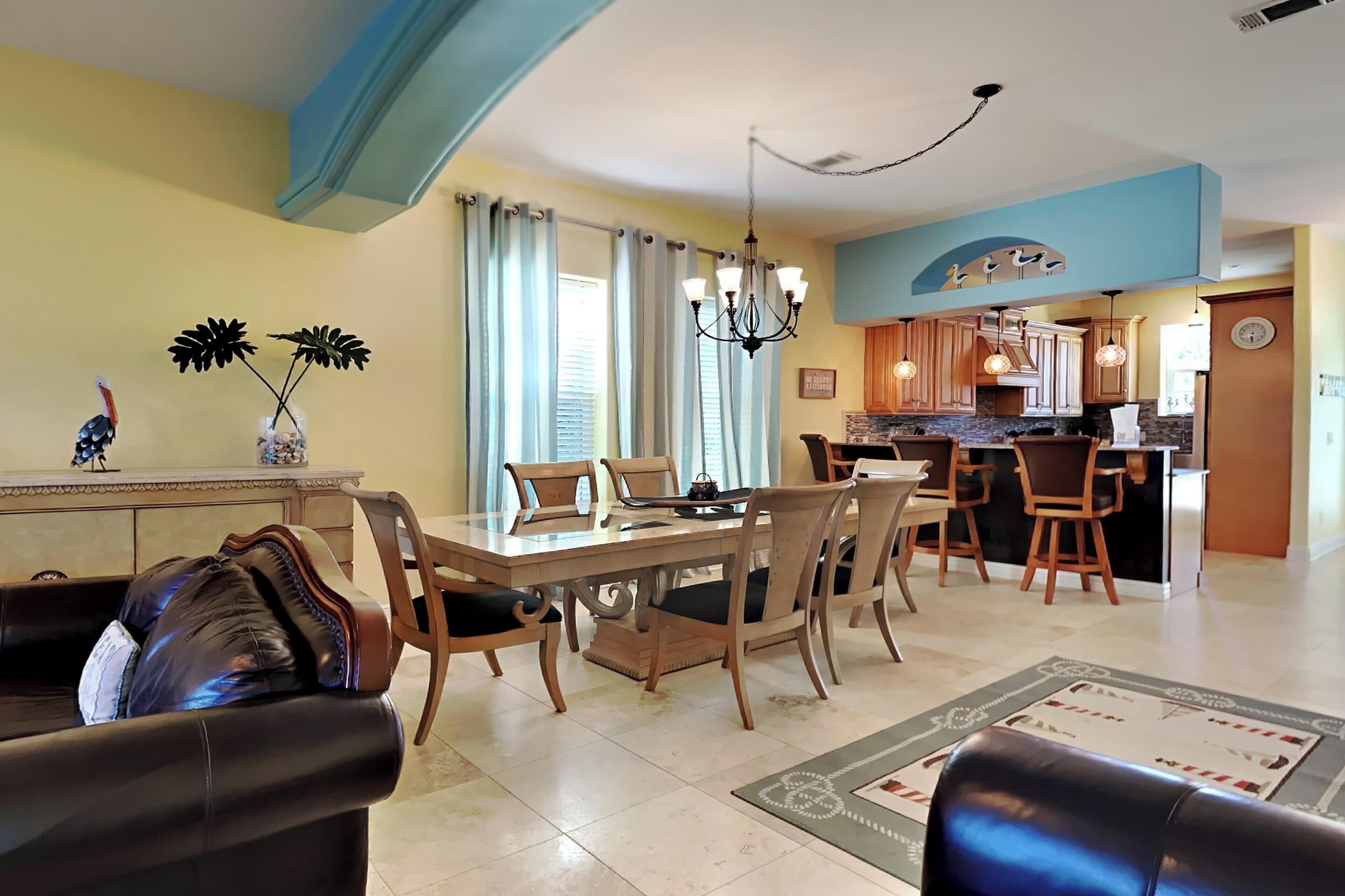 A dining area with a wooden table and chairs, a chandelier, and a kitchen in the background.