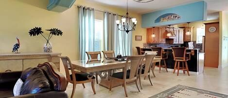 A dining area with a wooden table and chairs, a chandelier, and a kitchen in the background.