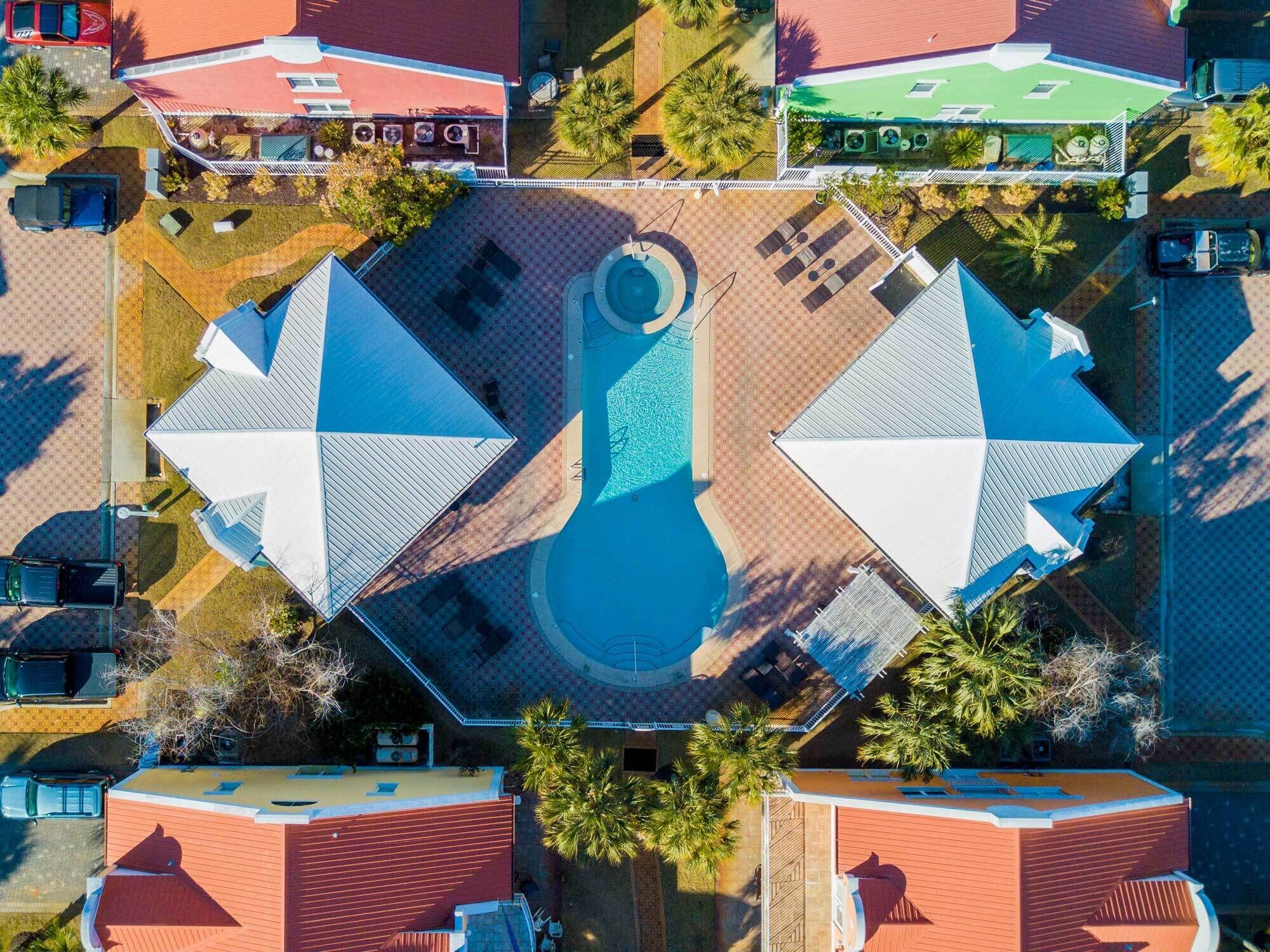 Vista aérea de uma propriedade com piscina central, cercada por um jardim com layout geométrico, e edifícios adjacentes com telhados vermelhos.