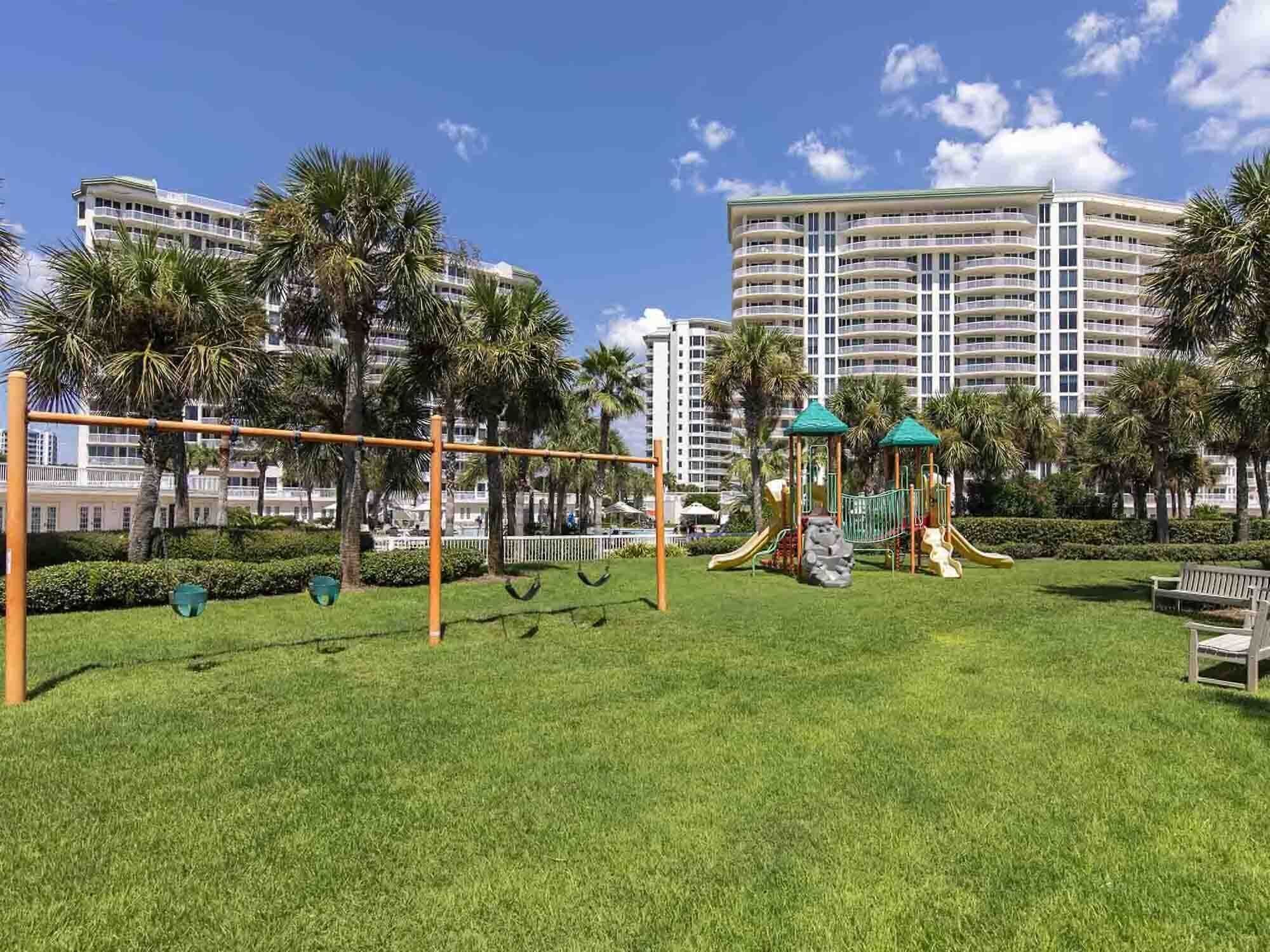 A playground with swings and slides, surrounded by well-maintained grass and palm trees, with high-rise buildings in the background.