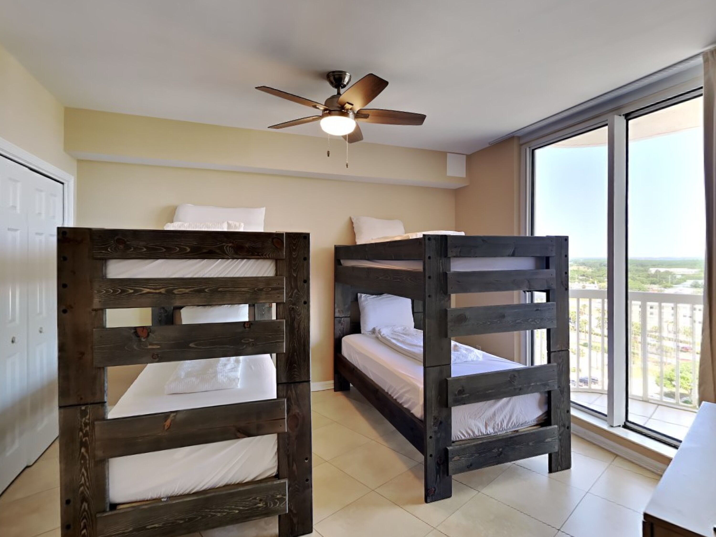 A room with two bunk beds, a ceiling fan, a large window with a view, and a white door.
