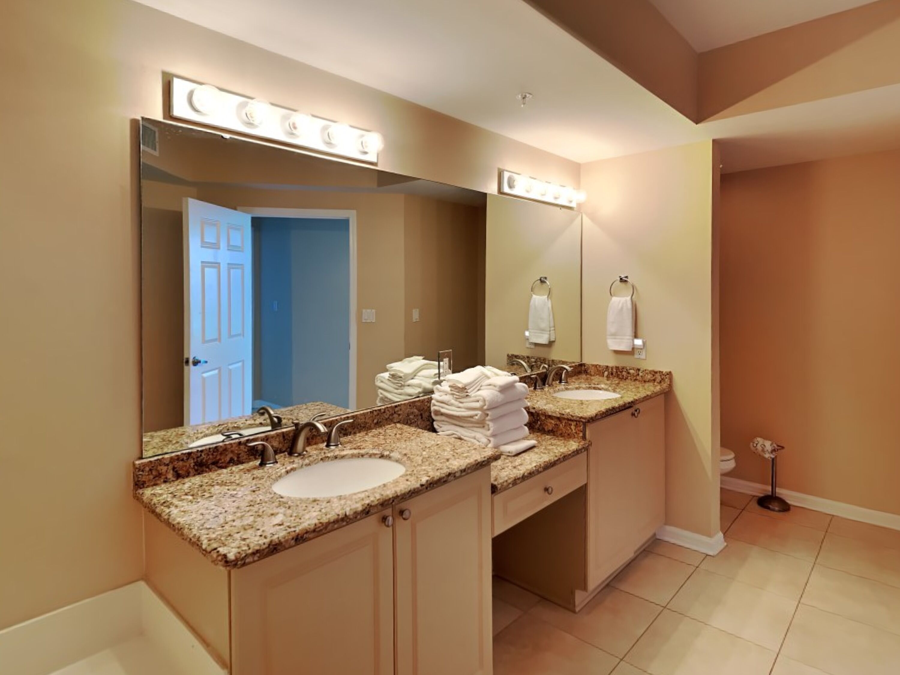 Une salle de bain moderne avec deux lavabos, un grand miroir avec éclairage intégré et une porte donnant sur une autre pièce.