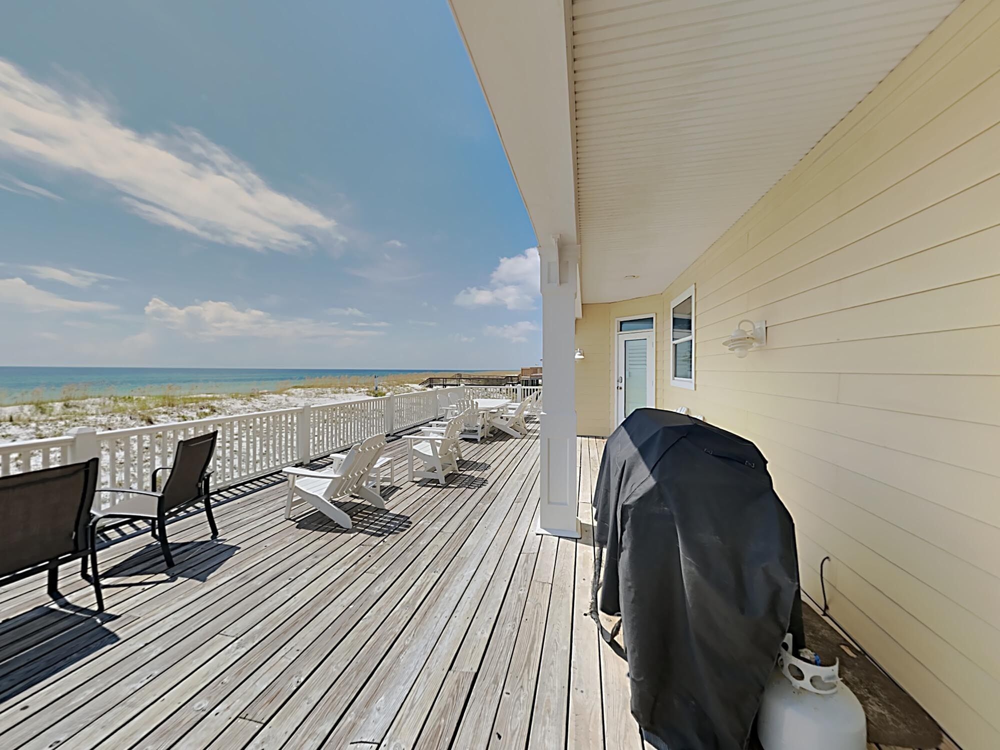 A wooden deck with white chairs and a barbecue grill, overlooking a beach.
