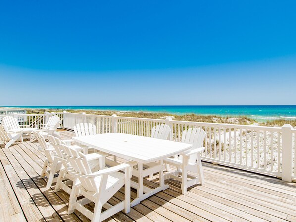 Una terraza de madera con sillas y una mesa blancos, con vista a una playa de aguas azules y una cerca blanca.