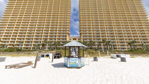 A large hotel with multiple floors and balconies, a beach area with sun loungers, and a sign for beach activities. - Calypso Resort & Towers #1809W by Resort Collection (Panama City Beach)