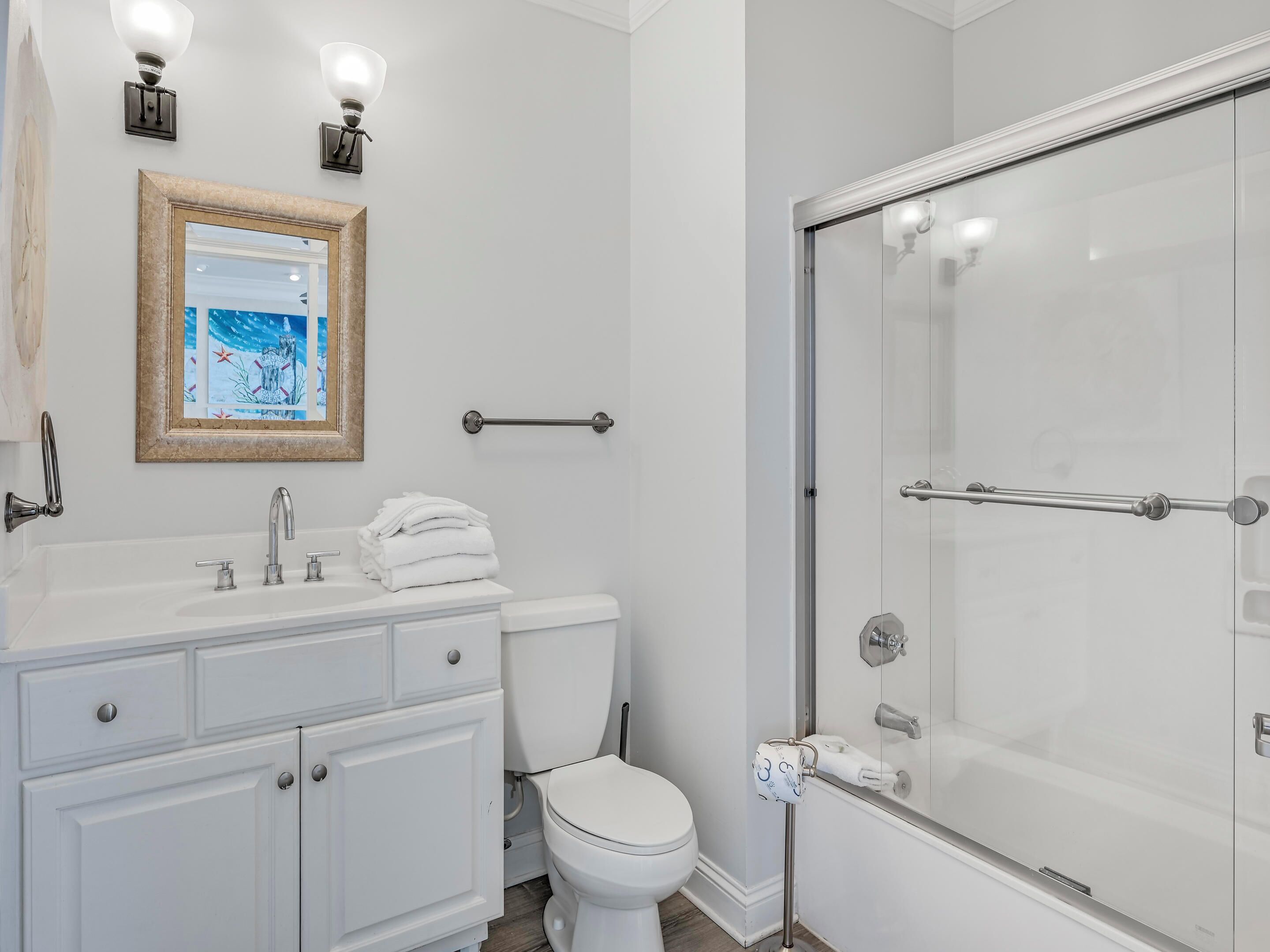 A modern bathroom with a white vanity, a framed artwork above the sink, a glass shower enclosure, and a toilet with a paper towel holder.
