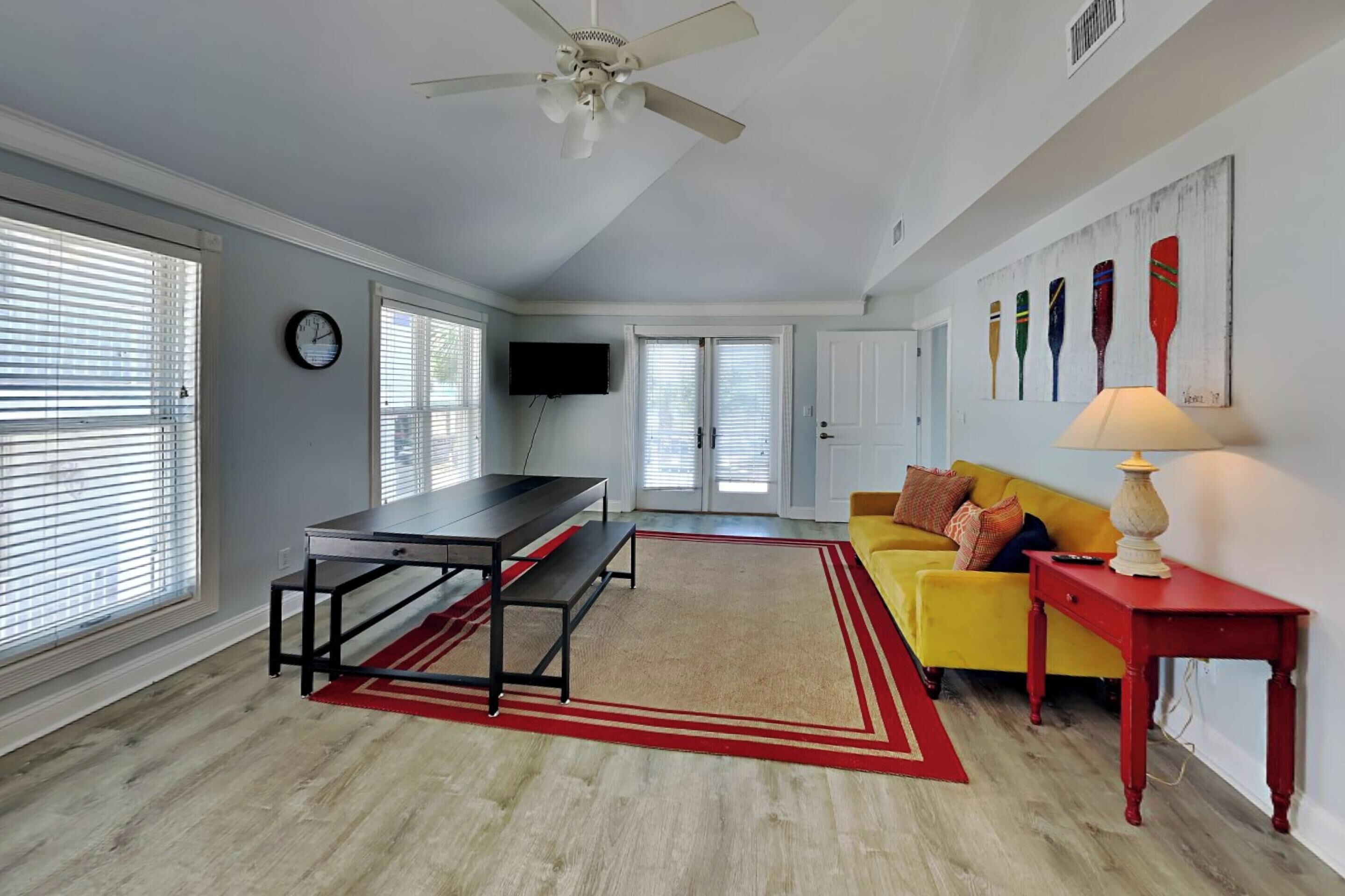Un salon spacieux avec un canapé jaune, une table rouge, un ventilateur de plafond et une grande fenêtre équipée de stores.