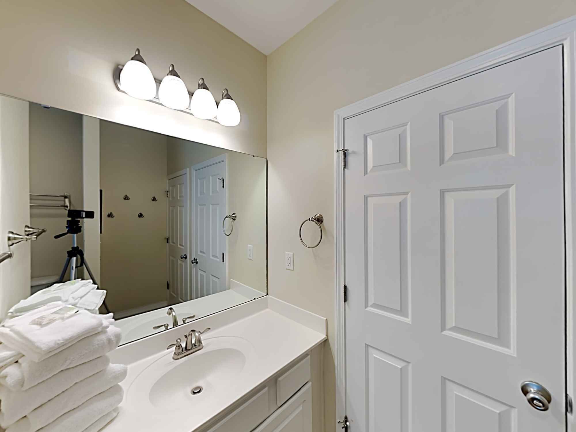 Une salle de bain avec un lavabo blanc, un miroir et une porte. Des serviettes sont pliées sur le lavabo et un appareil photo est installé sur un trépied dans un coin.