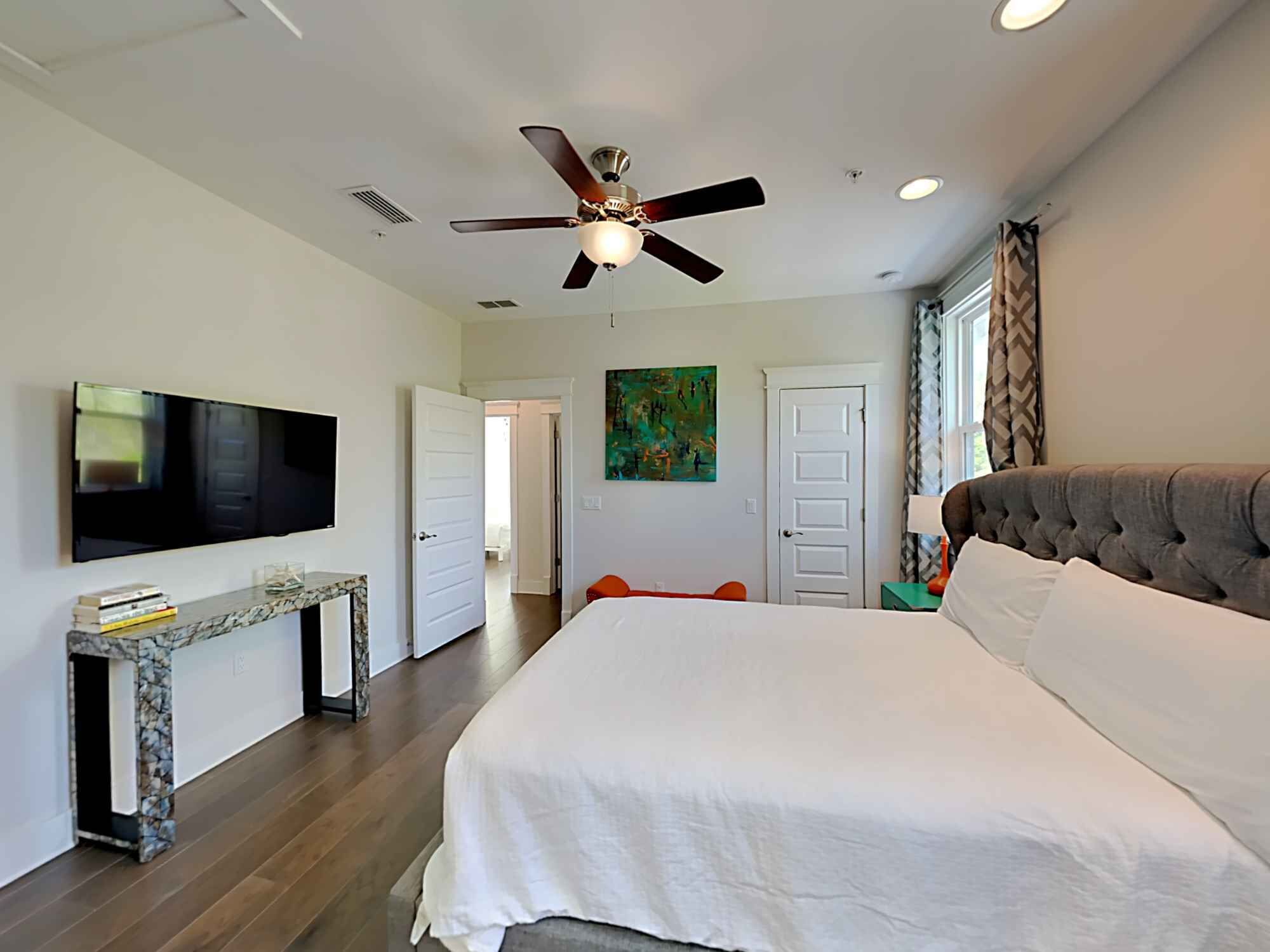 A bedroom with a large bed, a ceiling fan, a TV, and a painting on the wall.