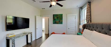 A bedroom with a large bed, a ceiling fan, a TV, and a painting on the wall.