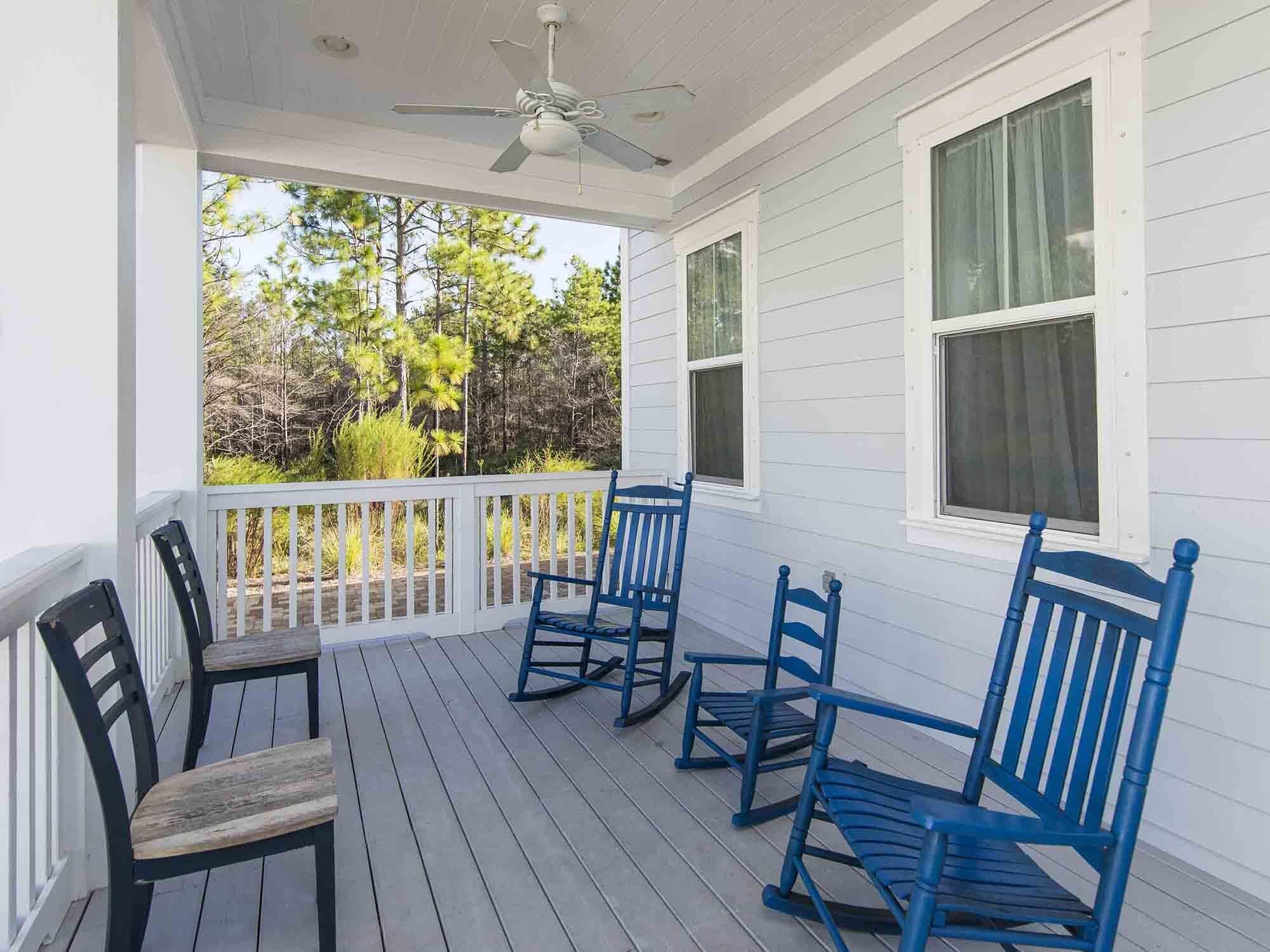 A covered porch with white railings, a ceiling fan, and a view of trees and shrubs.