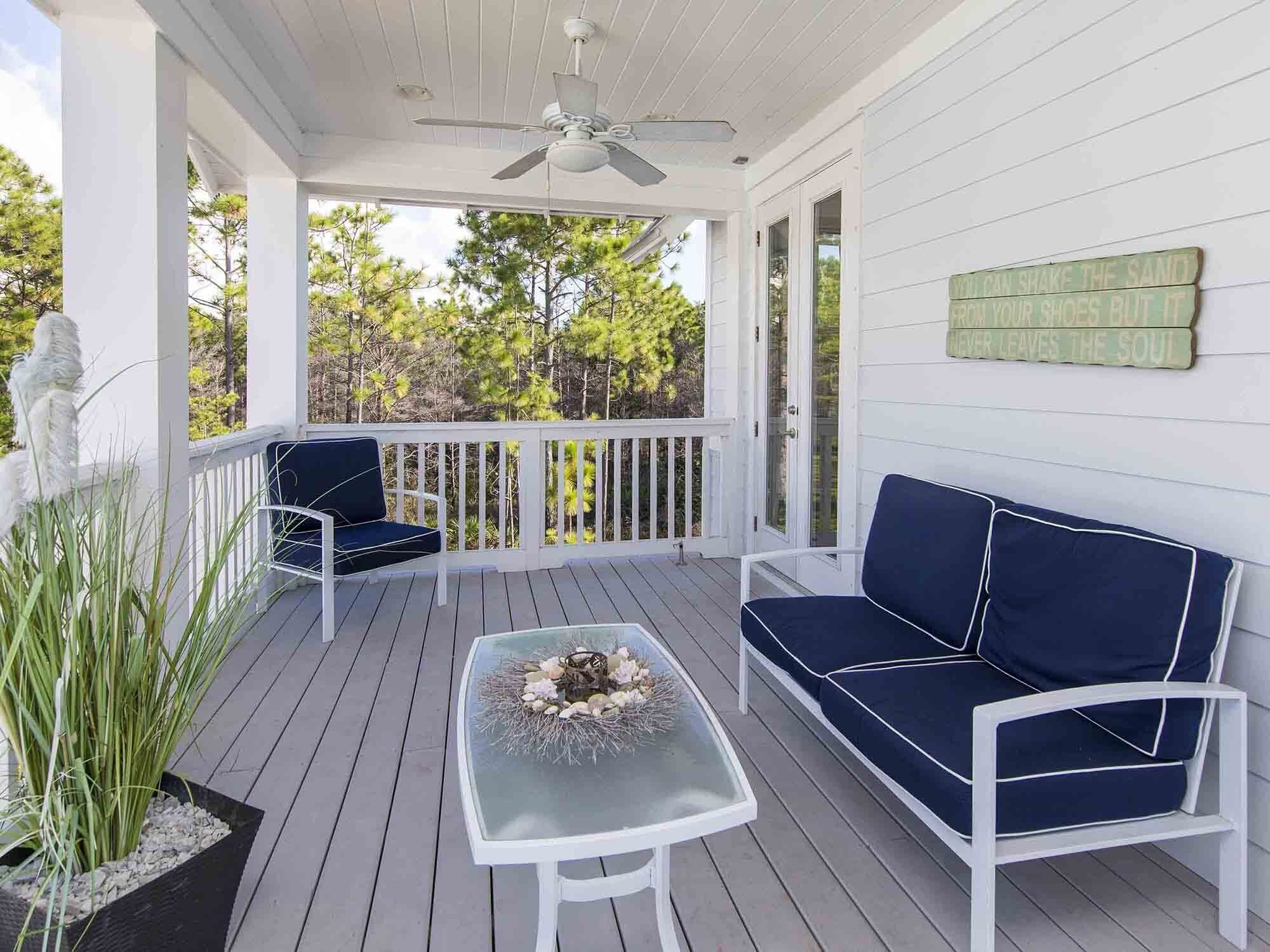 A covered porch with a ceiling fan, white railings, a dark blue sofa, a glass table with decorative items, and a potted plant.