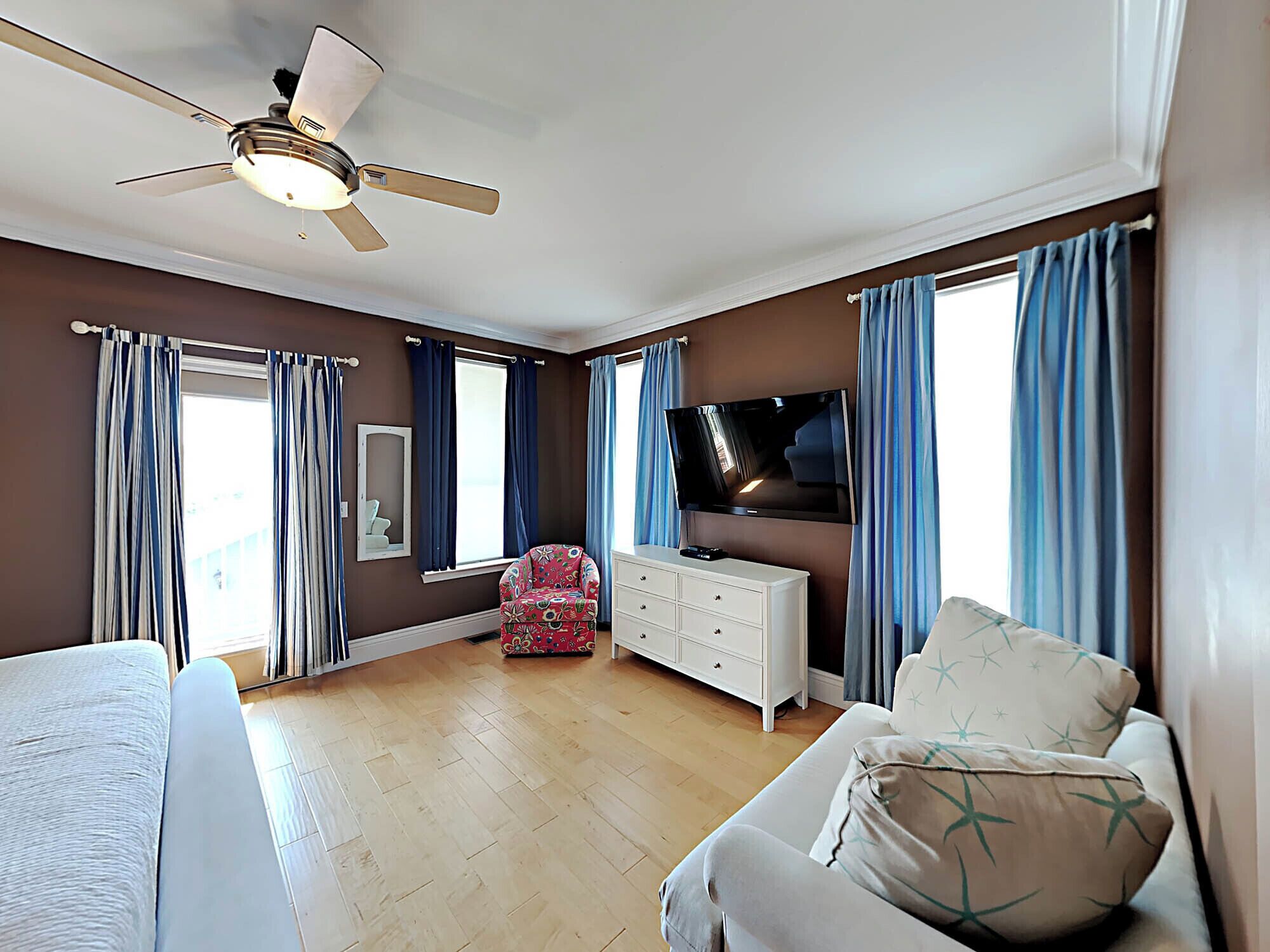 A bedroom with a ceiling fan, a television, a white dresser, a patterned armchair, and a sofa with star-patterned pillows.
