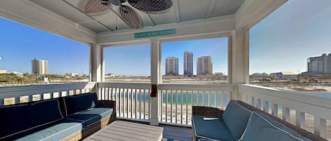 A covered outdoor seating area with wicker furniture, a ceiling fan, and a view of the beach and buildings.
