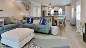 Ein geräumiges Wohnzimmer mit Sofa, Hocker und einem Essbereich, im Hintergrund ist eine Küche zu sehen.