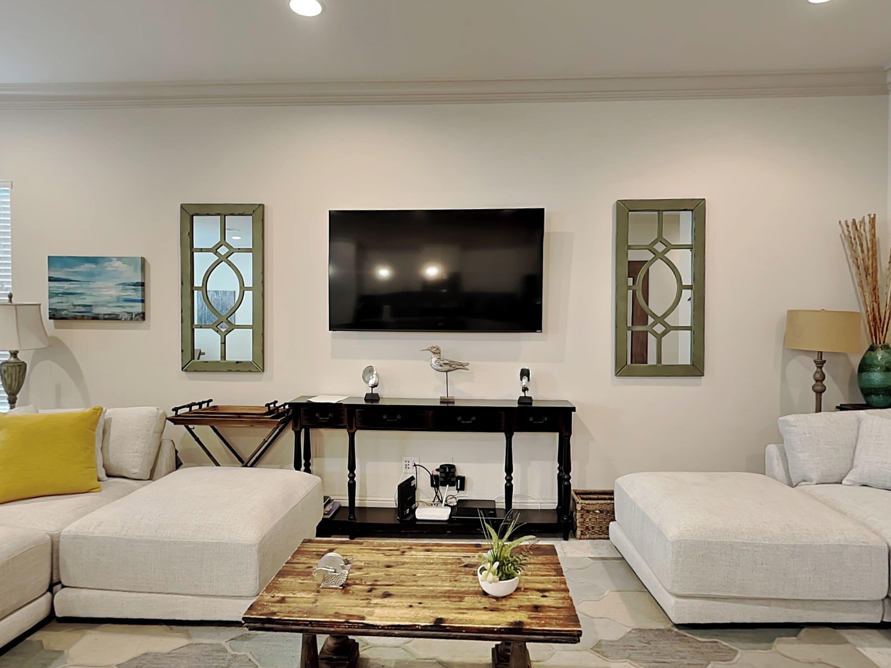 A living room with a sofa set, a coffee table with decorative items, a television mounted on the wall, and two decorative mirrors.